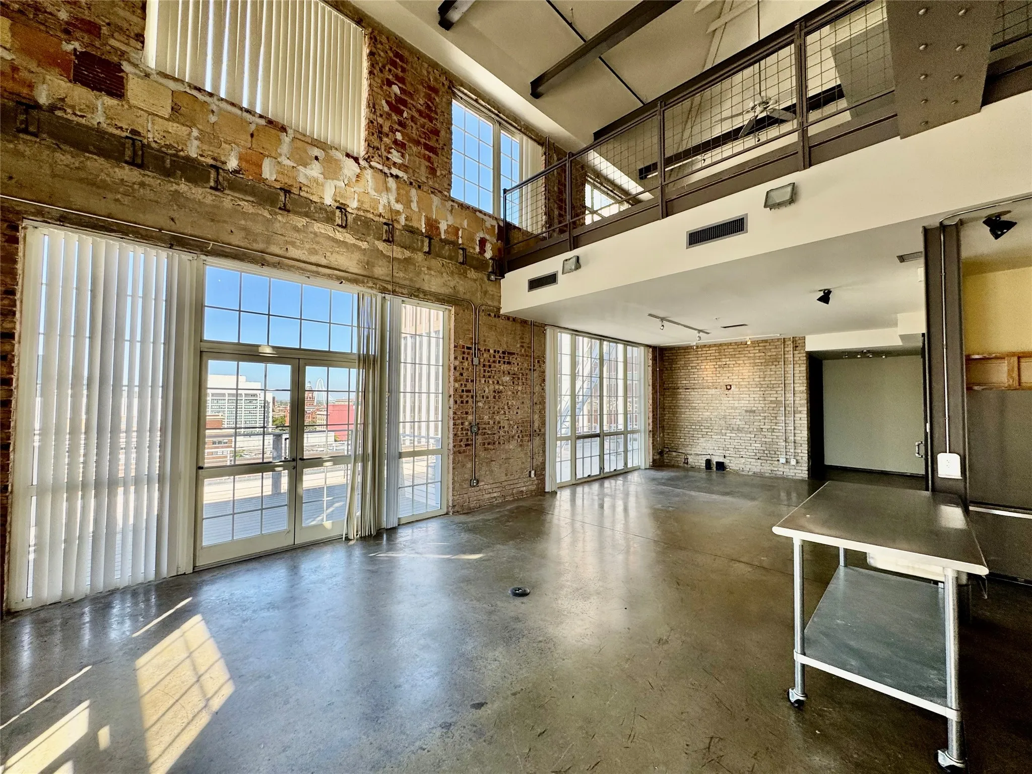 Exposed brick walls, concrete flooring, large grid-patterned windows, high ceilings, and an open metal railing mezzanine