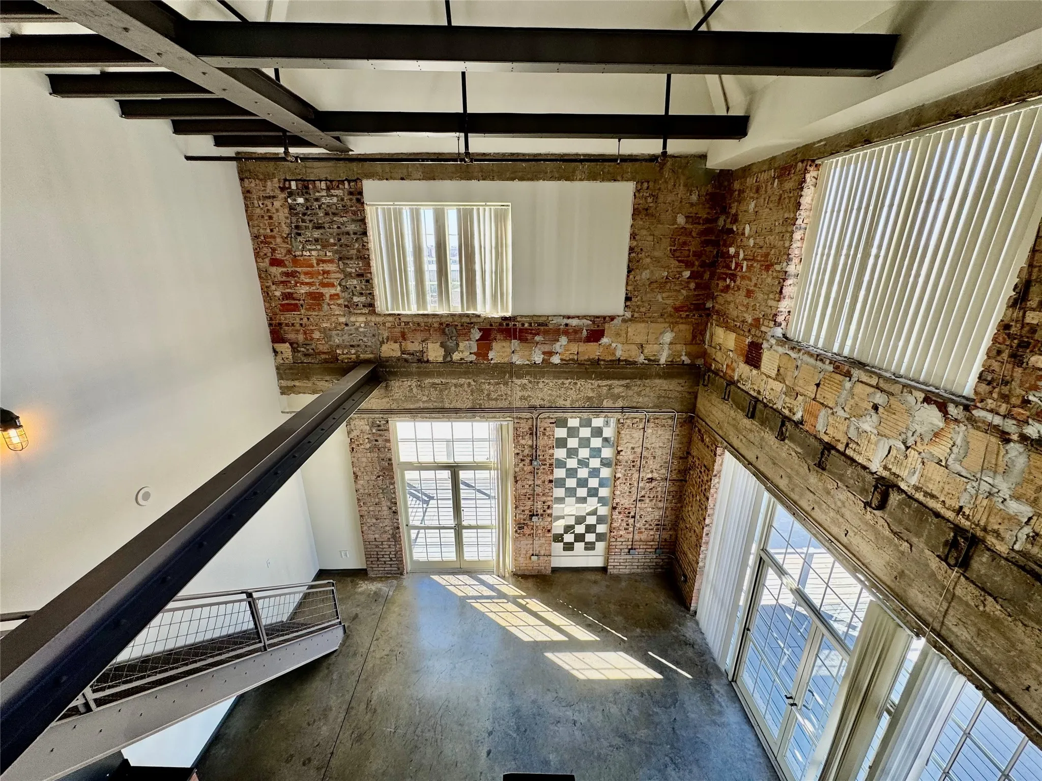 Exposed brick walls, concrete flooring, large industrial-style windows, open metal ceiling beams, and a metal-railed staircase