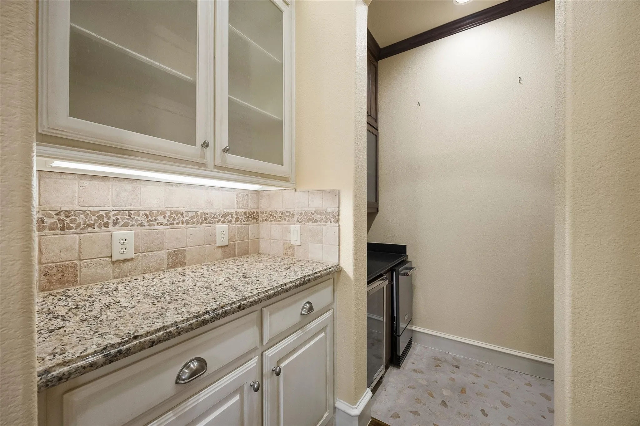 White cabinetry with frosted glass inserts, speckled granite-style countertop, mosaic and square tile backsplash, integrated under-cabinet lighting, and stone tile flooring