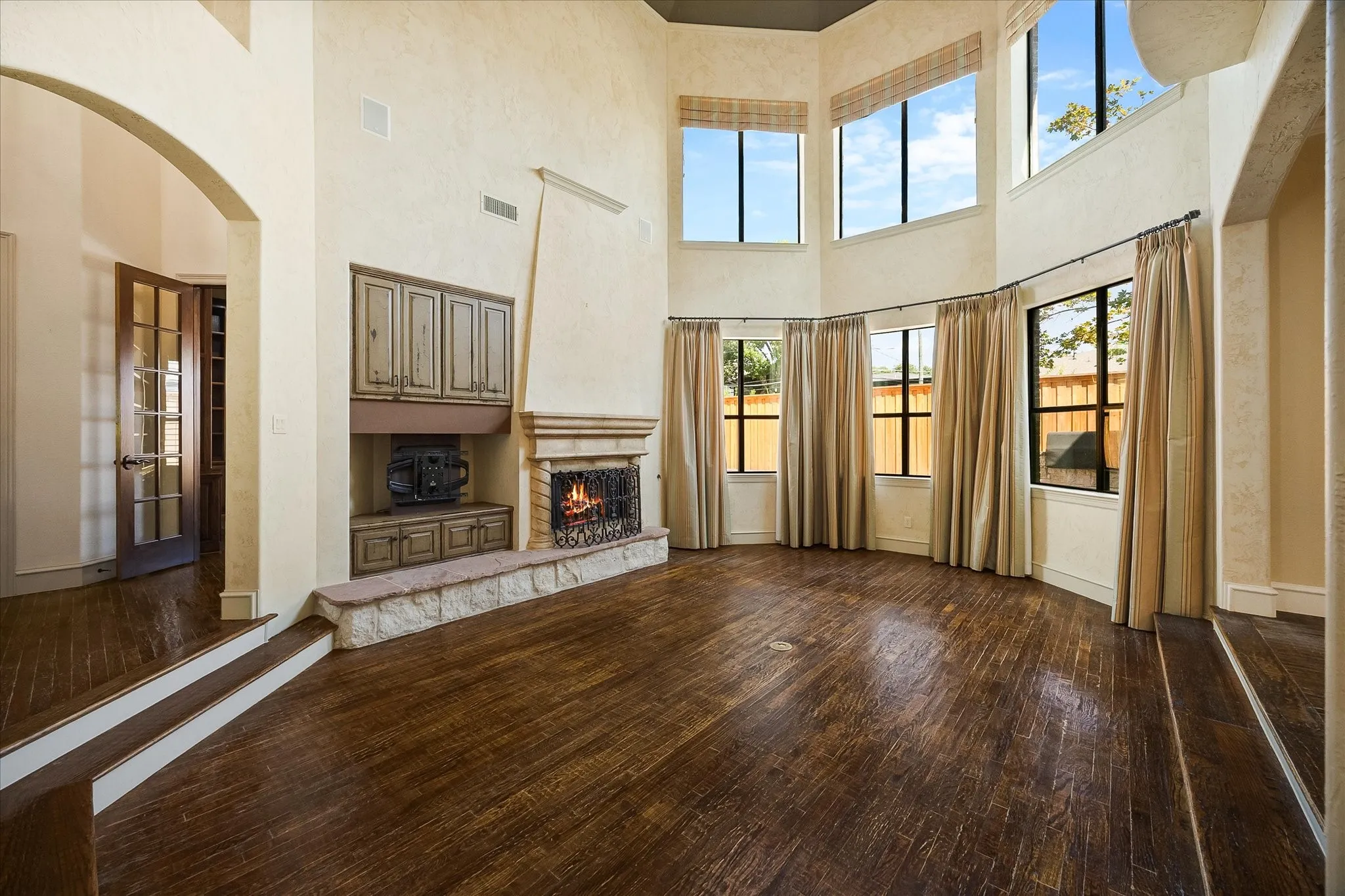 Spacious living area featuring high ceilings, wood-finish flooring, a stone fireplace with hearth, built-in cabinetry, and large windows