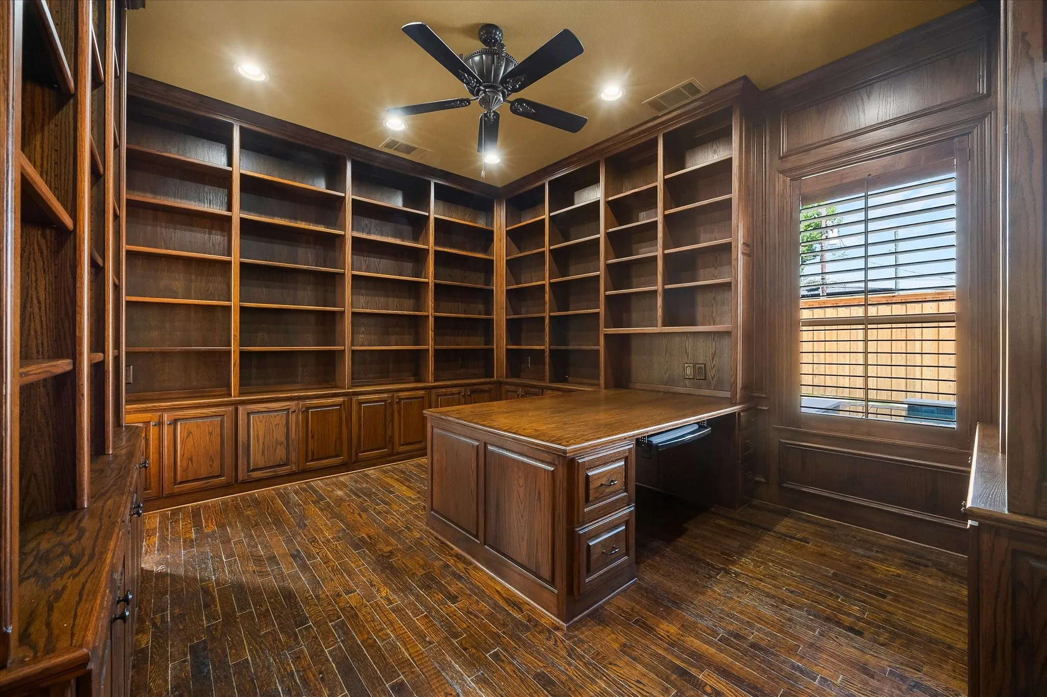 Extensive built-in shelving and cabinetry, rich wood-finish flooring, integrated desk, ceiling fan, and window with plantation shutters