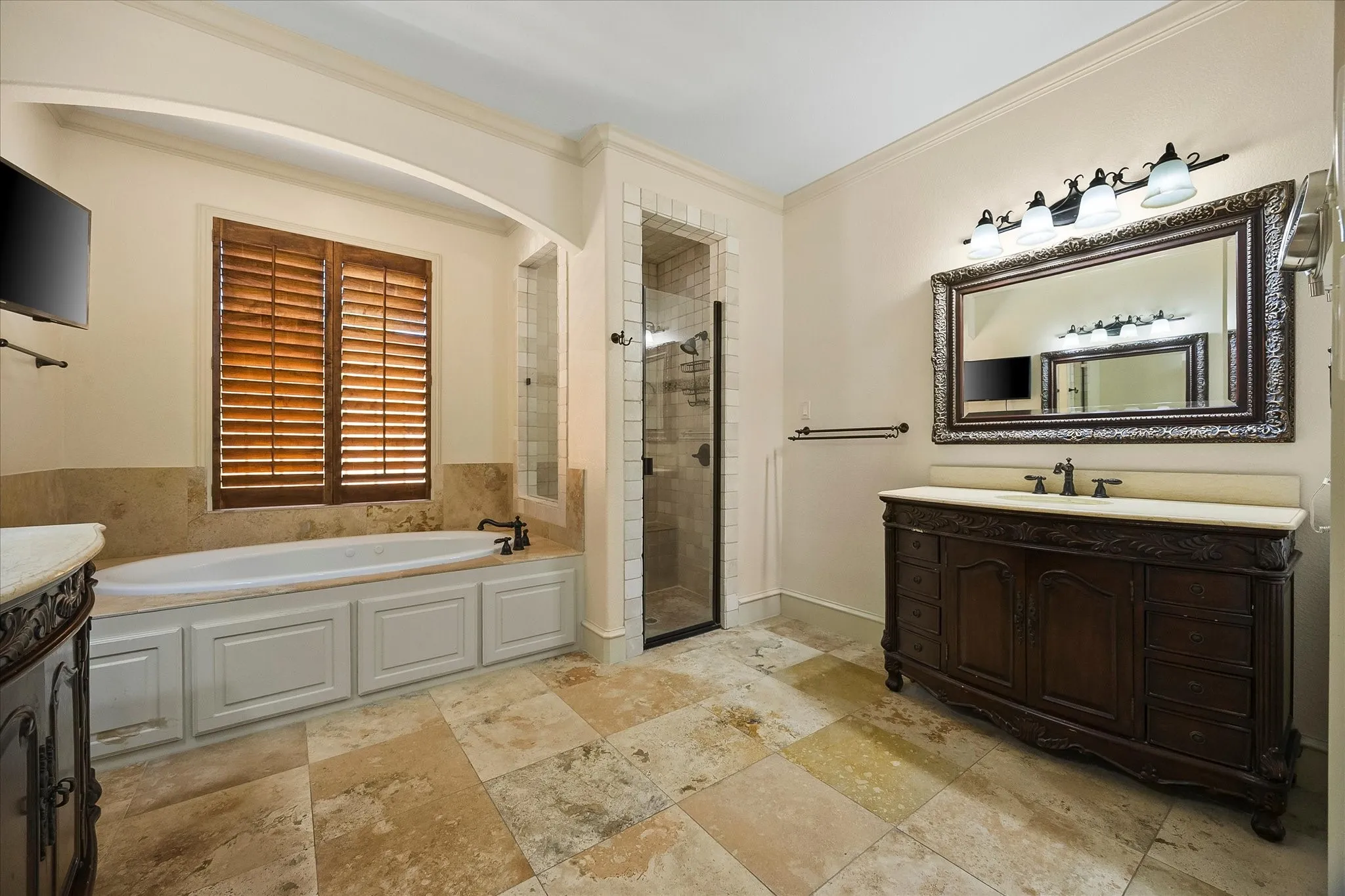 Built-in soaking tub with wood-toned shutters, walk-in shower with glass enclosure, ornate dark wood vanity with counter, and large decorative mirror with overhead lighting