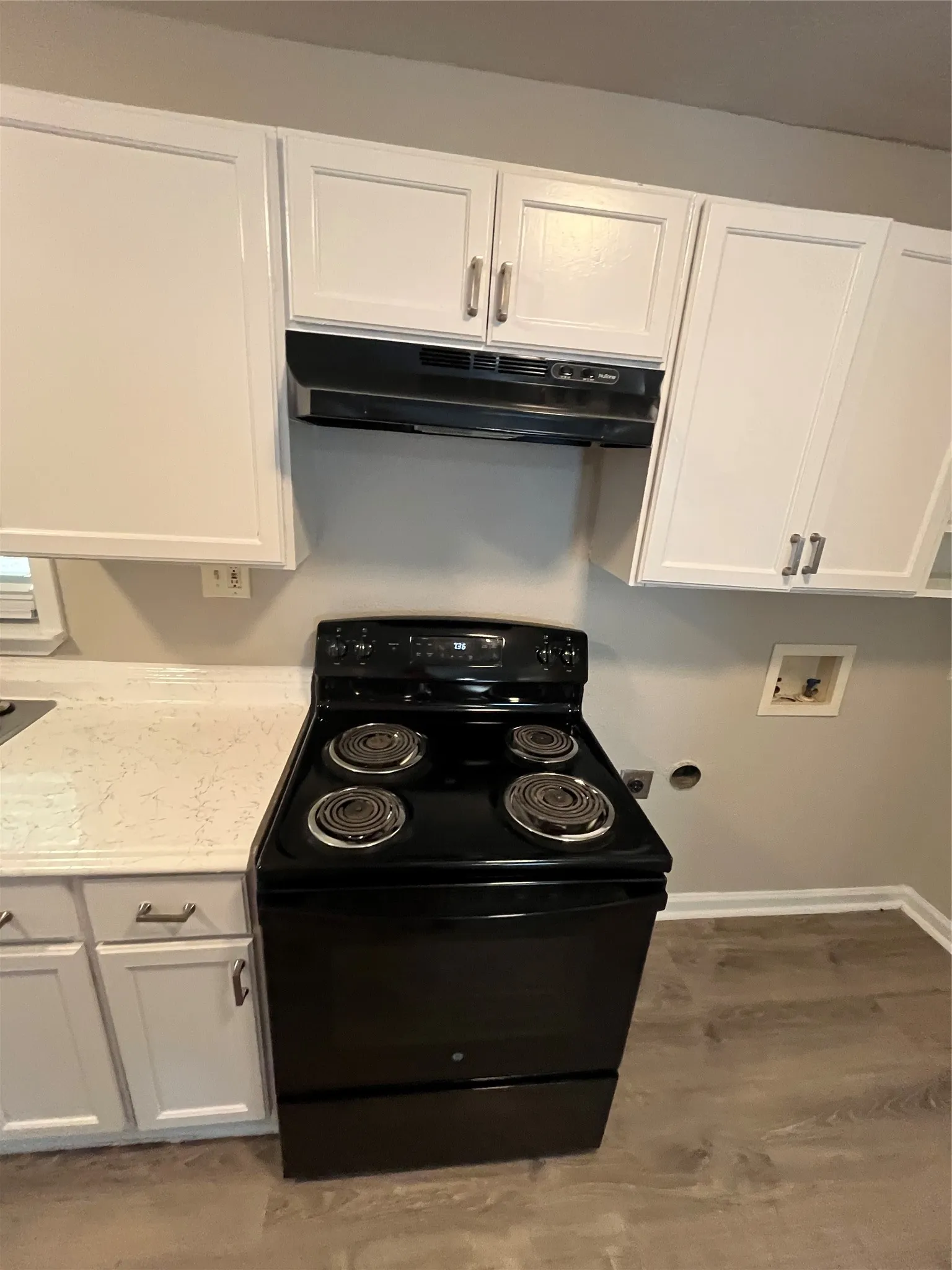 White shaker-style cabinetry with silver-tone hardware, black electric range with coordinating range hood, light-toned countertop, and wood-finish flooring