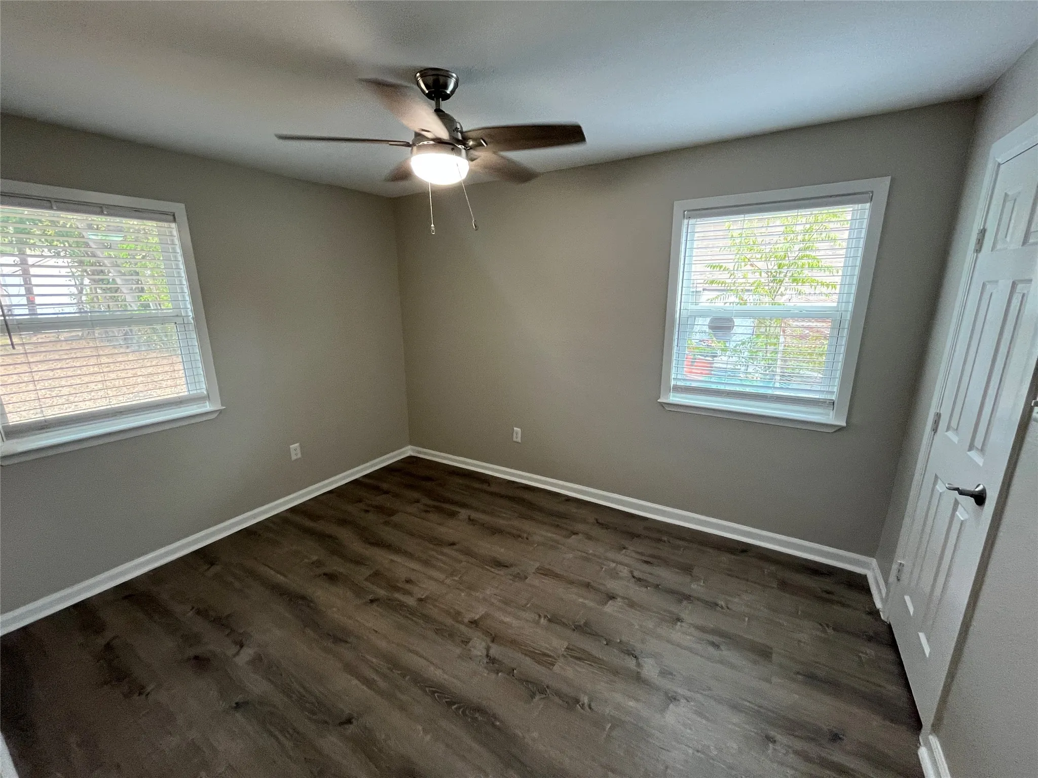 Neutral-toned walls, wood-finish flooring, two windows with blinds, a ceiling fan with integrated lighting, and a white paneled door