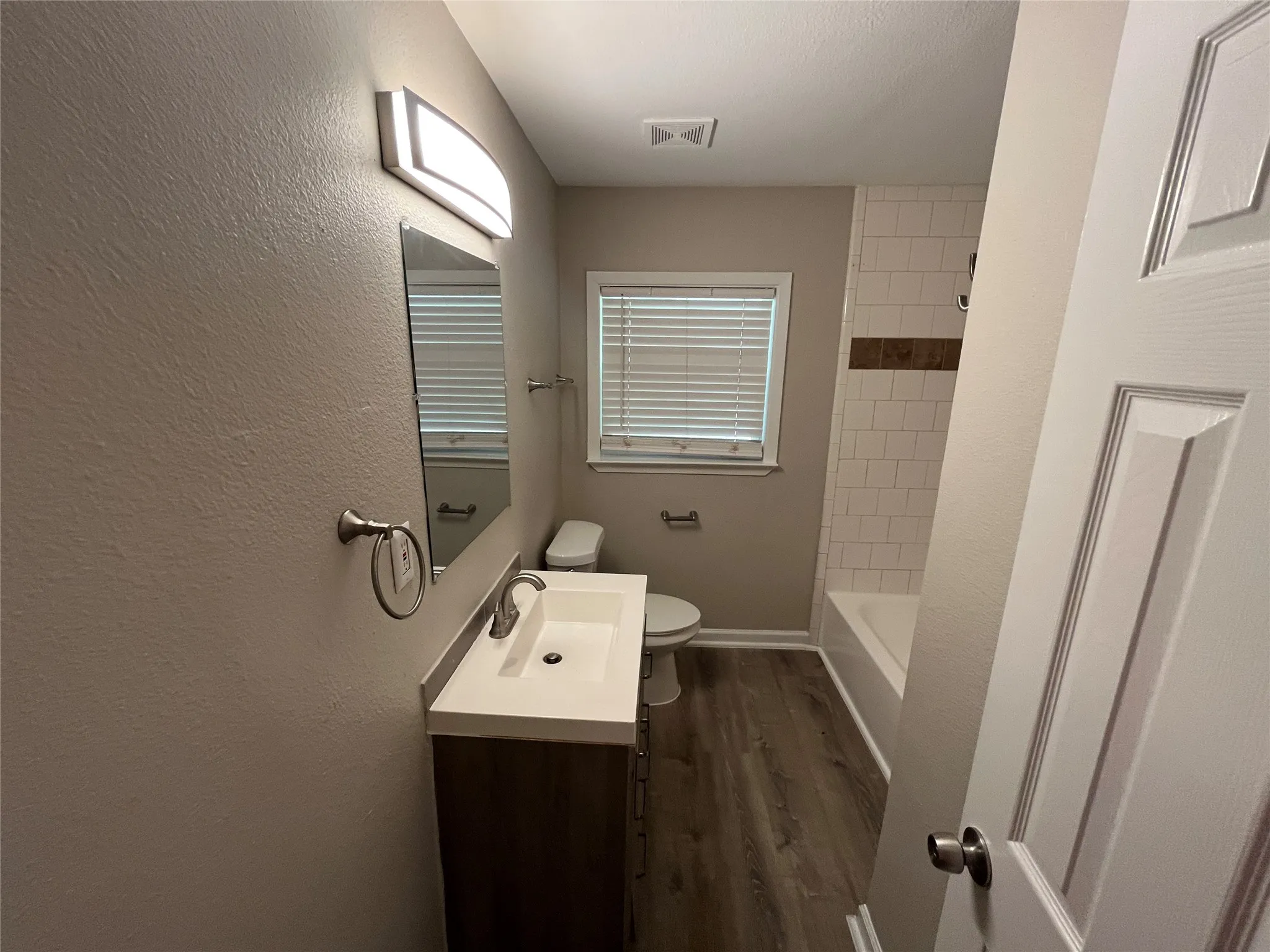 Bathroom featuring a single vanity, wood-finish flooring, a full bathtub with subway tile surround, and a window with blinds