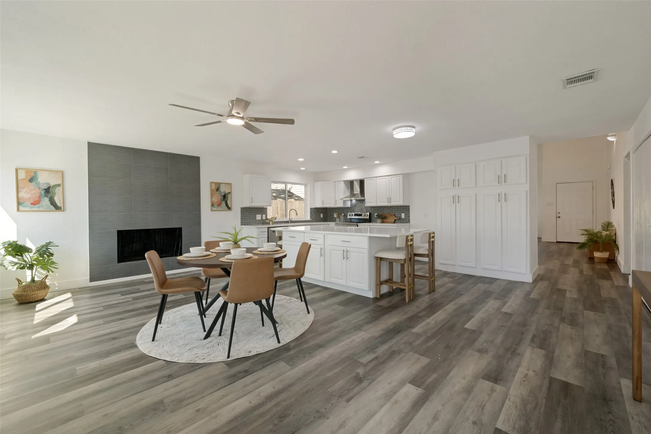 Open-concept living space featuring wood-finish flooring, a modern fireplace with dark tile surround, a kitchen island with seating, white shaker cabinetry, and a ceiling fan