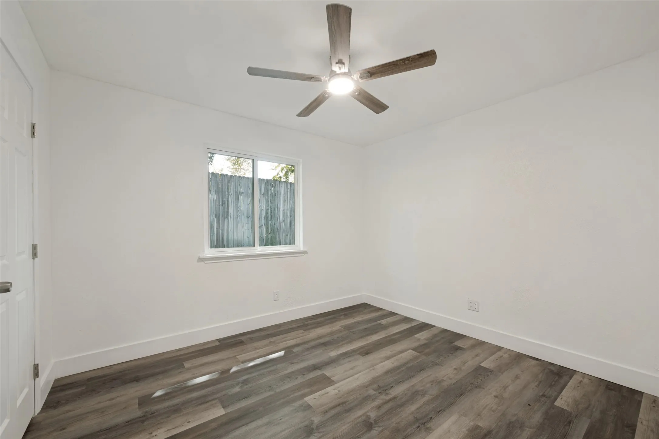 Wood-finish flooring, white walls, ceiling-mounted fan with integrated lighting, and a white-framed window