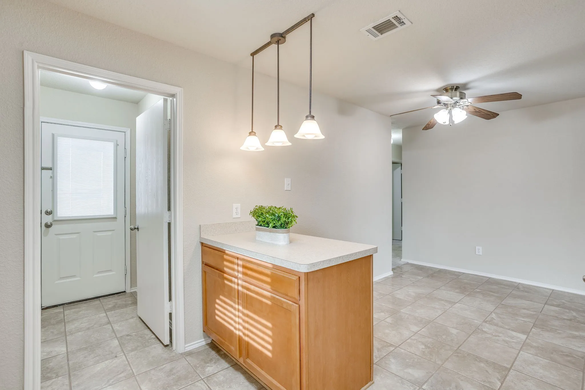Light-toned tile flooring, island with wood-finish cabinetry, drop pendant lighting, ceiling fan, and a white exterior door