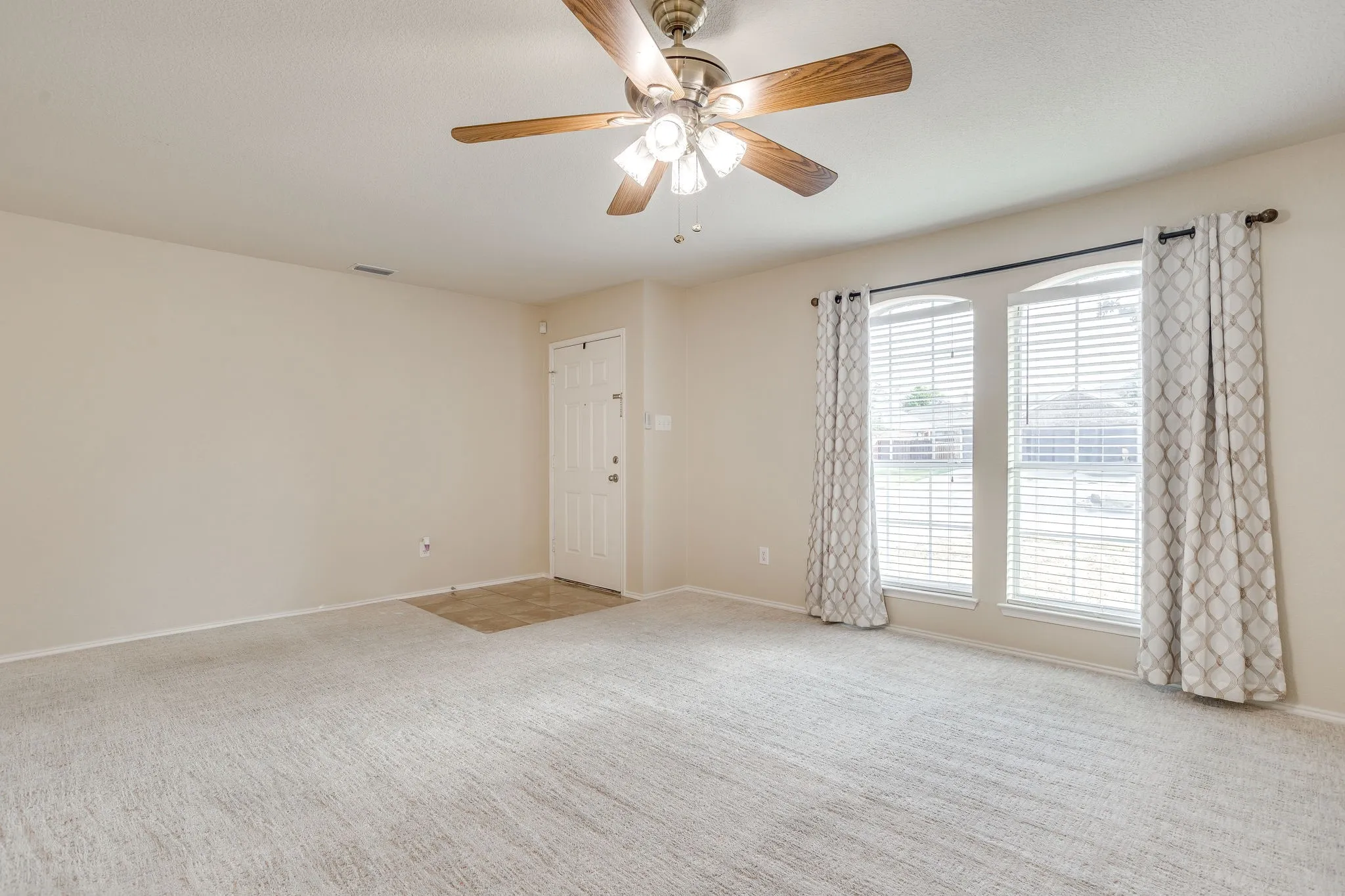 Spacious room with light-colored carpeting, a ceiling fan with light fixture, and a front entry door with a tile-finish landing