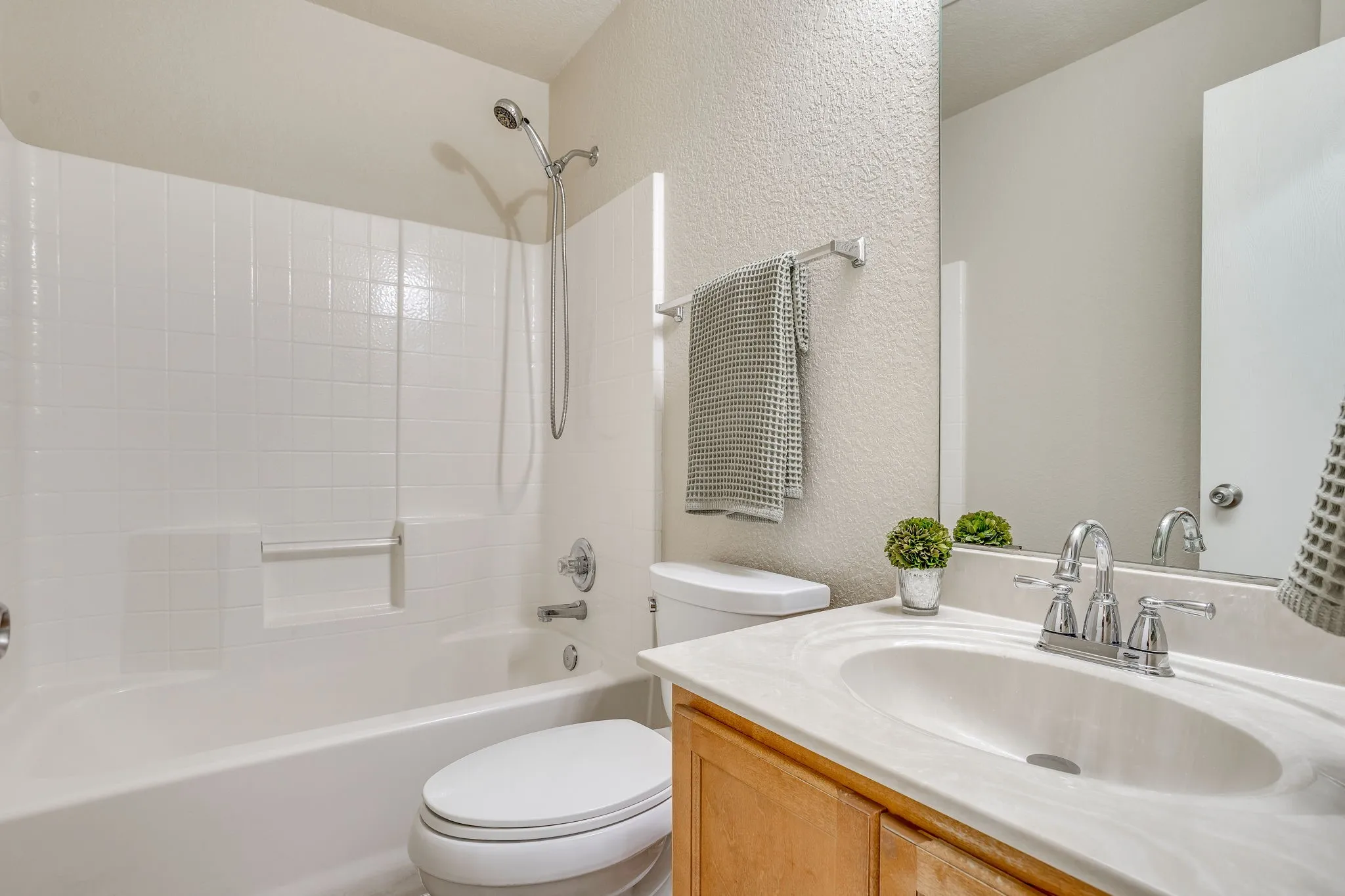 White tiled shower/tub combination with a handheld shower head, wood-finish vanity with an integrated sink, and chrome fixtures