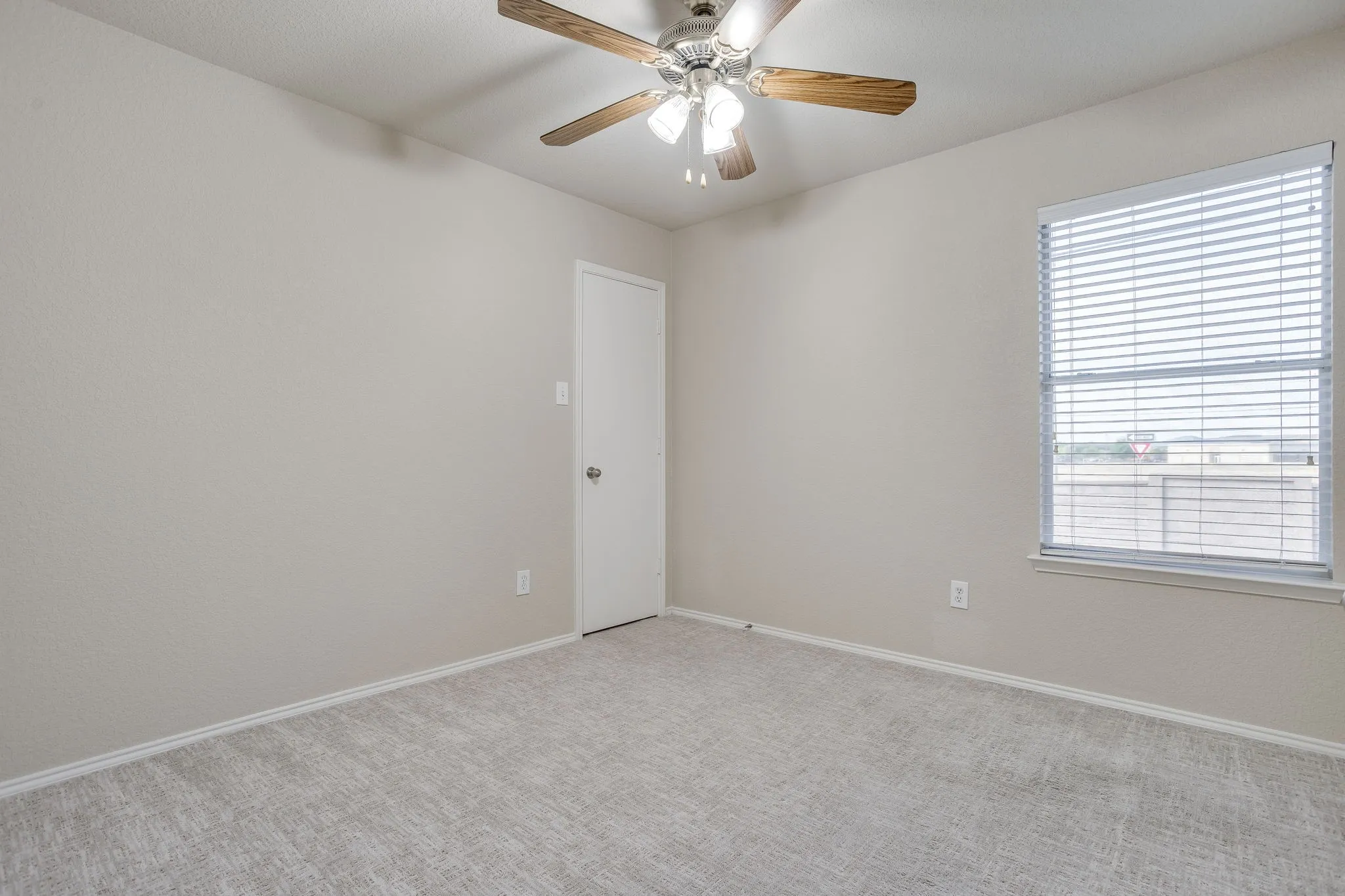 Neutral tone wall finish, light gray carpet flooring, ceiling fan with wood-finish blades, single window with blinds, and white interior door