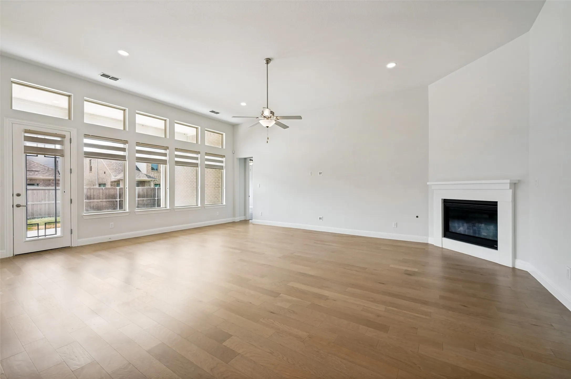 Expansive and light-filled living room featuring soaring ceilings, beautiful hardwood floors, and a wall of windows that fills the space with natural light. A cozy fireplace creates an inviting focal point, while the spacious open layout offers plenty of room for comfortable seating and entertaining.