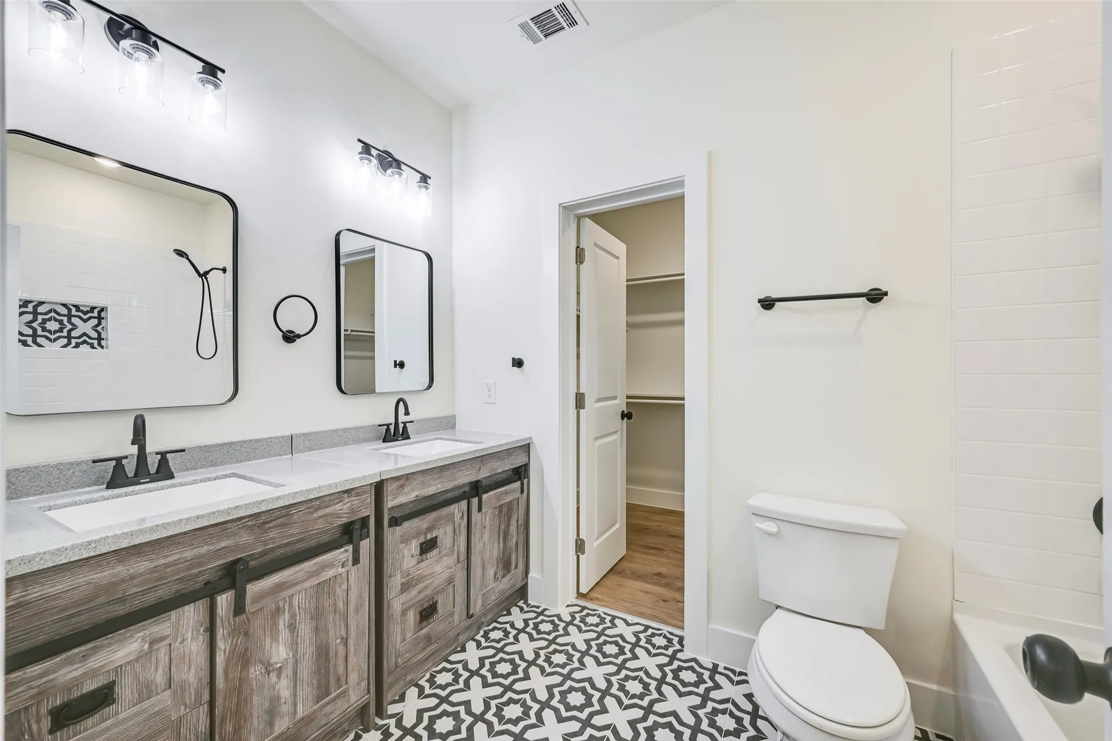 Bathroom featuring a dual vanity with light-toned countertops, wood-finish cabinetry with black hardware, and patterned floor tiling