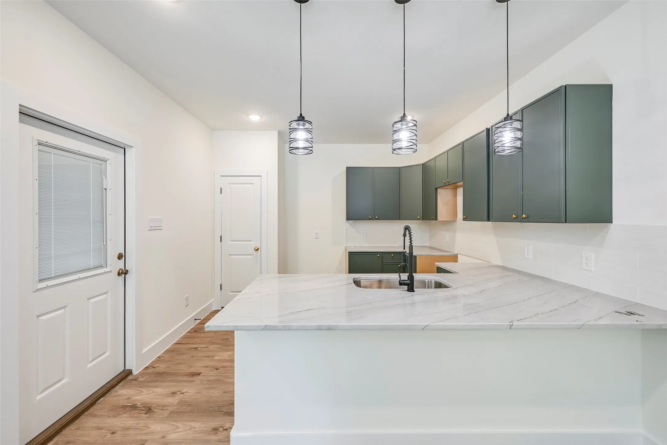 Kitchen featuring a white quartz-finish island with an undermount sink and matte black faucet, dark green cabinetry, white subway tile backsplash, and wood-finish flooring