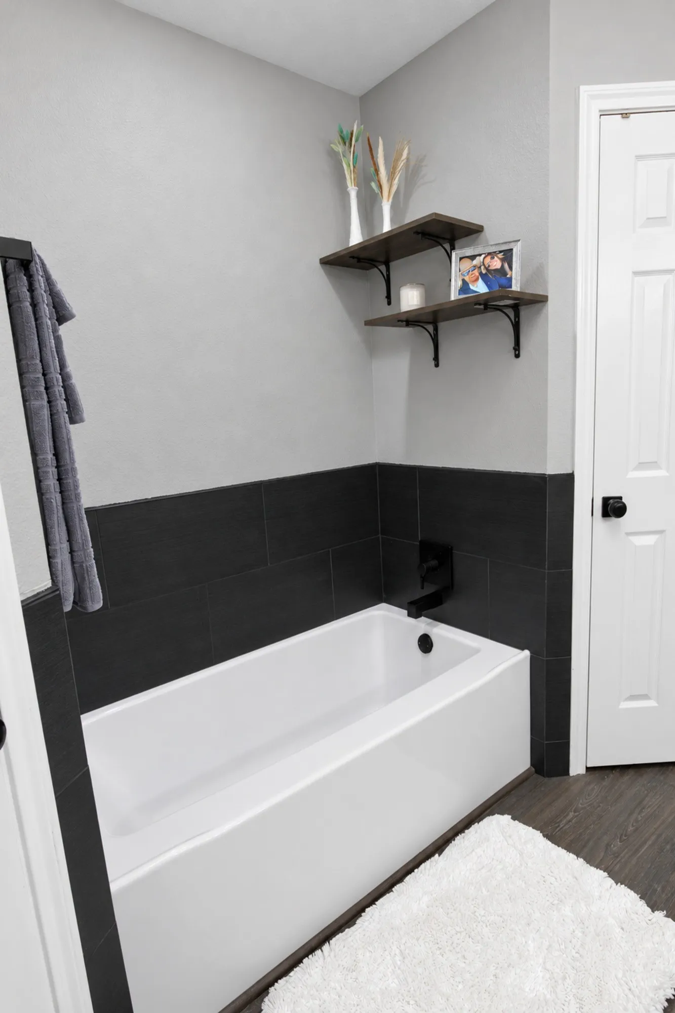 White bathtub with matte black fixtures, dark tile surround, and wood-finish flooring