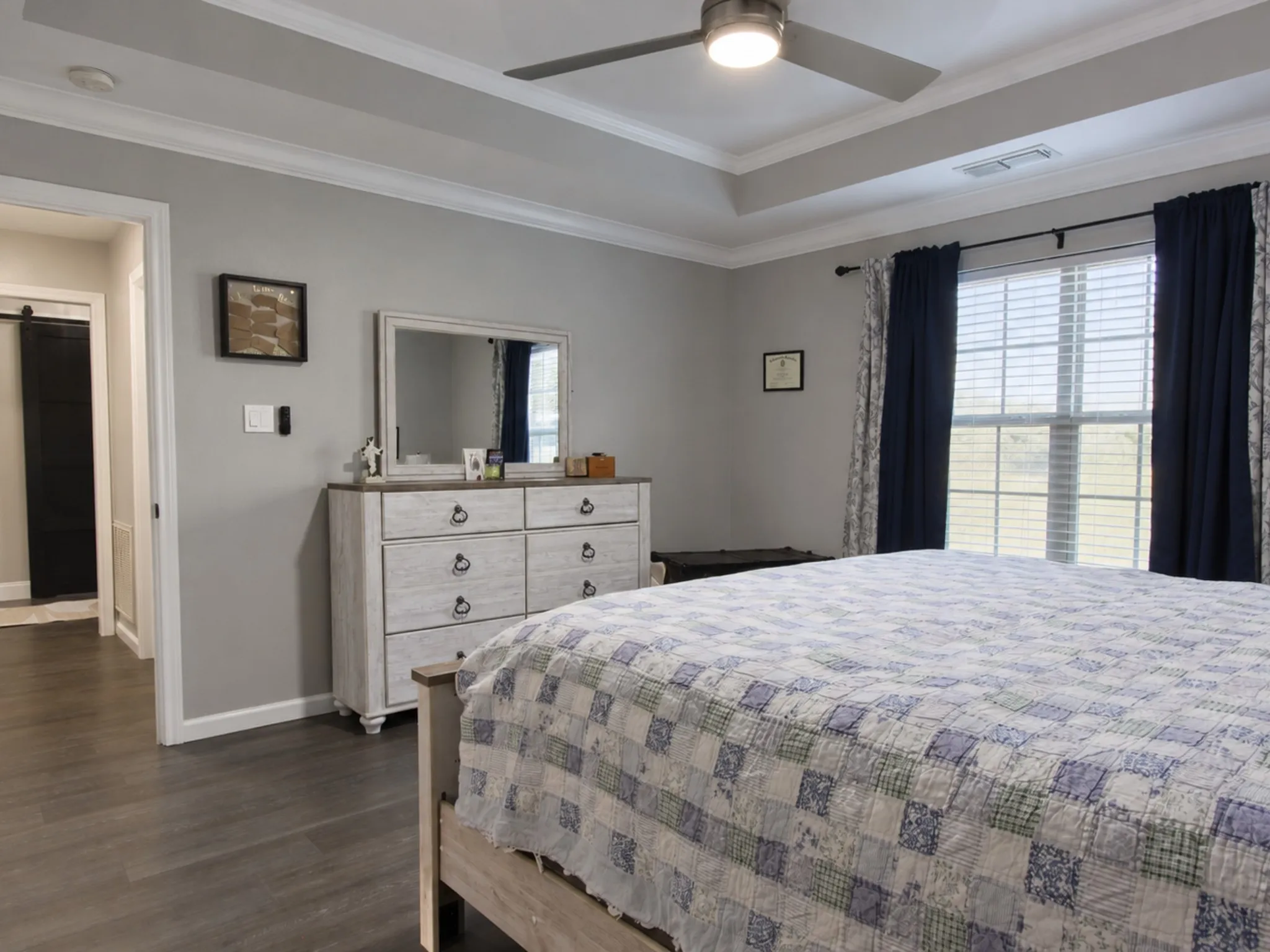 Tray ceiling with crown molding, ceiling fan with integrated lighting, window with blinds, wood-finish flooring, and a barn door entry to an adjoining space