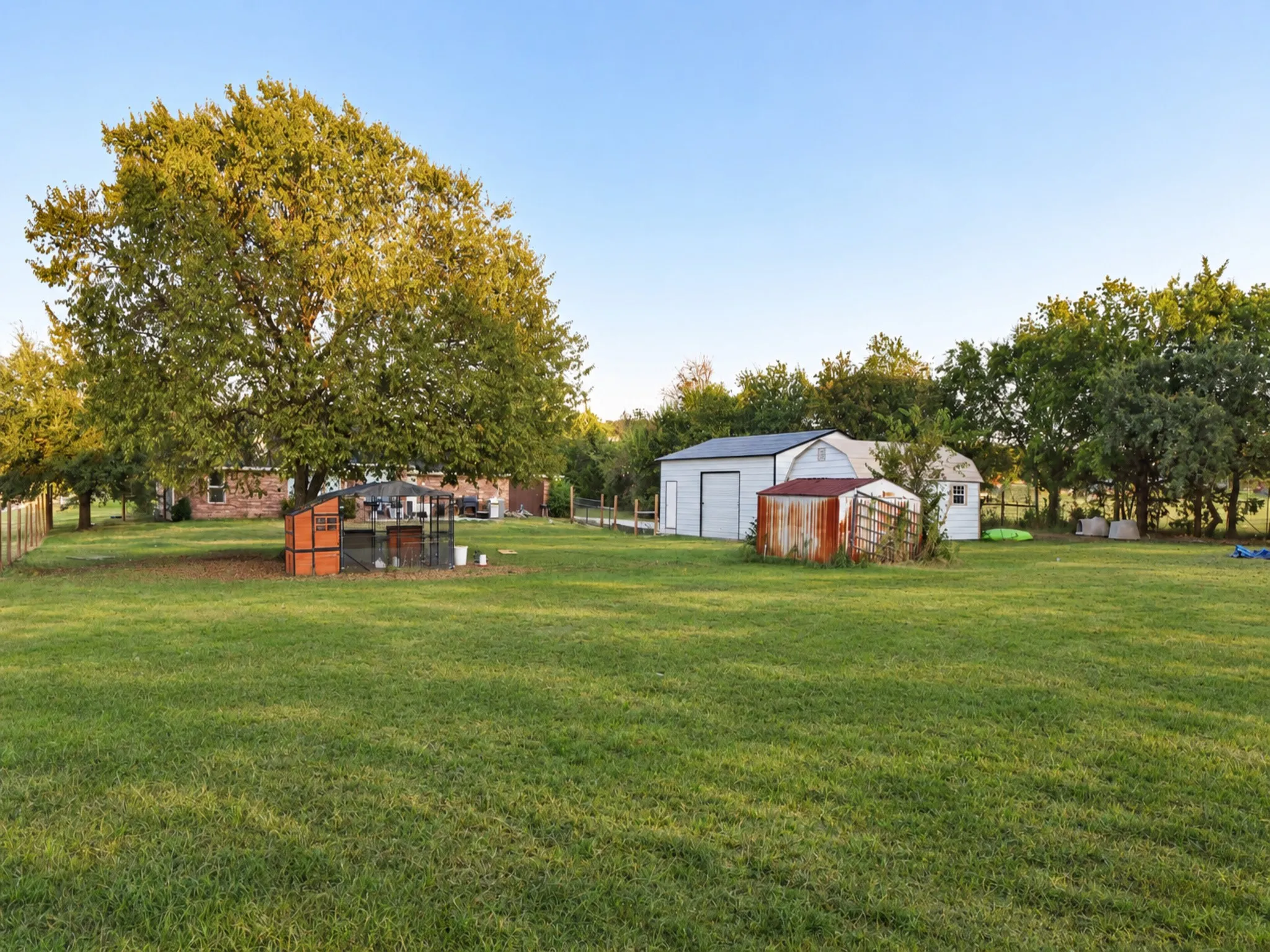 Expansive green lawn with mature trees, featuring a white detached garage, a white shed, and a fenced area