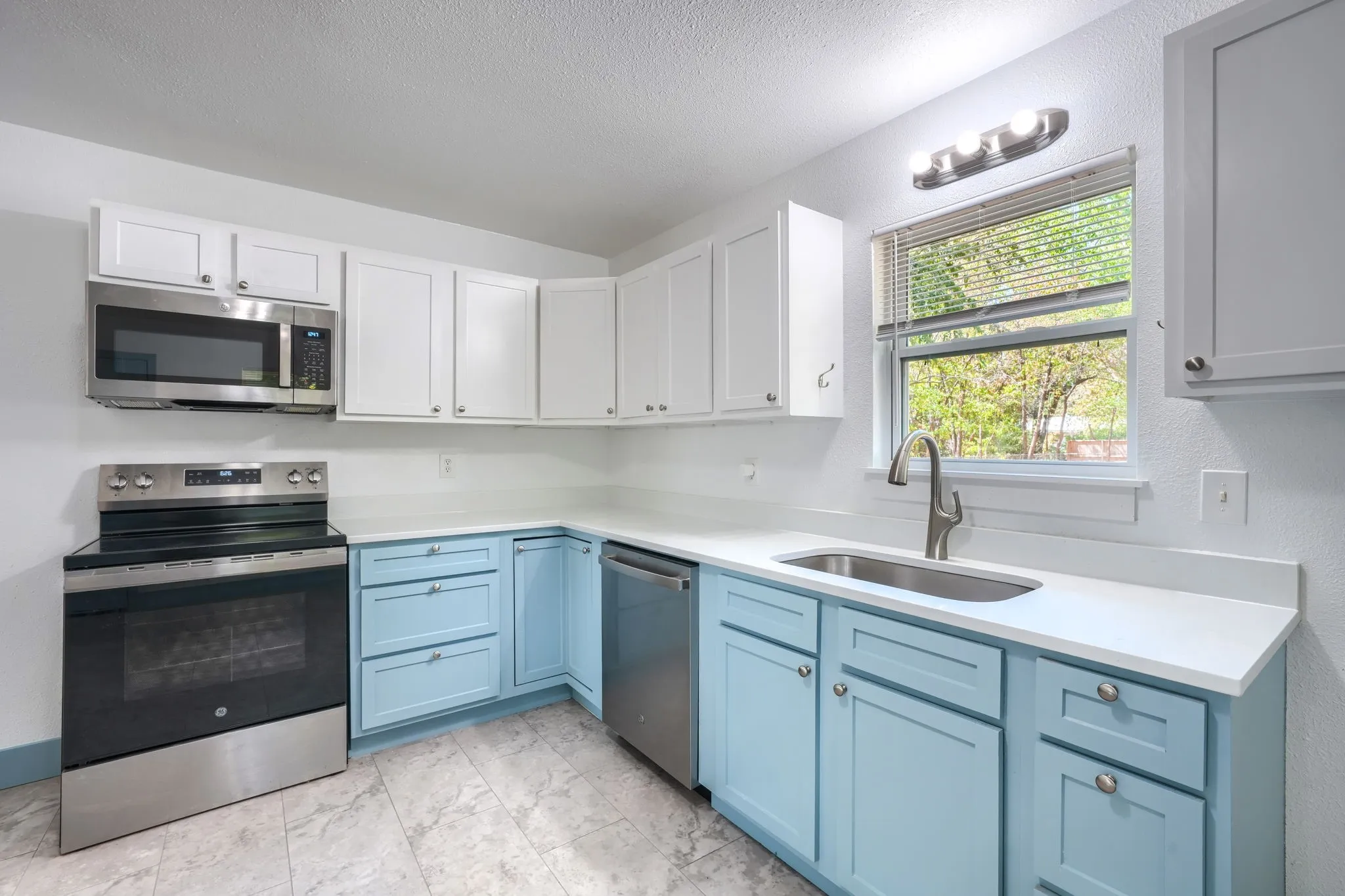 Kitchen featuring white upper cabinetry, blue lower cabinetry, stainless steel appliances, a single basin sink with a gooseneck faucet, and light-colored countertops