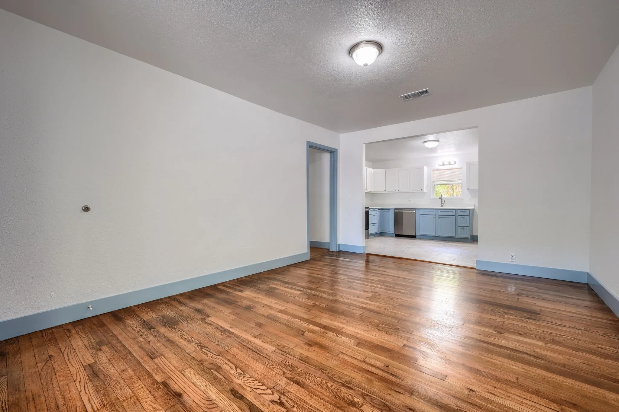 Hardwood flooring throughout, white walls, and a recessed kitchen with light blue lower cabinetry and stainless steel appliances
