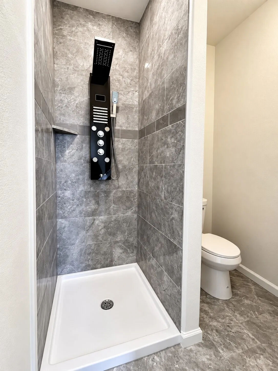 Shower featuring gray marbled wall tile, a dark-toned shower panel with multiple spray options, and a built-in shower niche