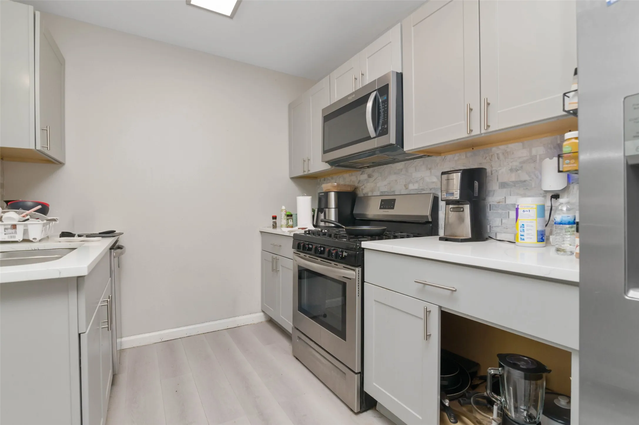 Galley kitchen featuring light gray cabinetry, white countertops, tile backsplash, wood-finish flooring, and stainless steel appliances including a range and microwave