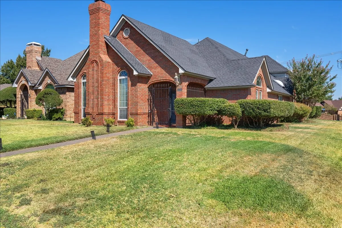 Brick exterior with a prominent chimney, arched windows, dark shingle roofing, and established shrubbery