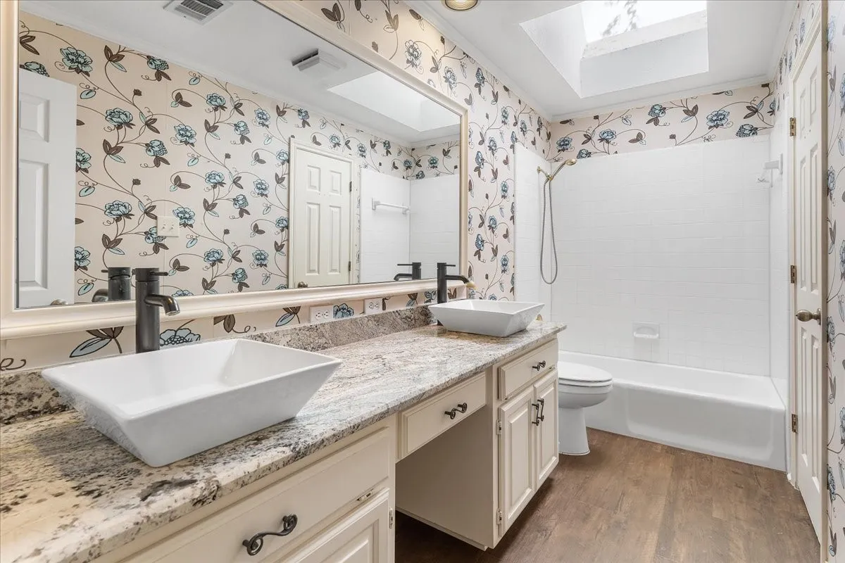Skylight, wood-finish flooring, white vanity cabinetry with dark hardware, dual vessel sinks with dark faucets, and a bathtub with shower