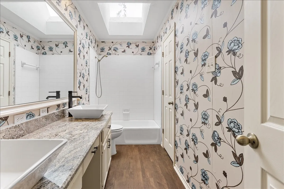 Dual vessel sinks, stone-finish countertop, wood-finish flooring, full-size bathtub with shower, and overhead skylights
