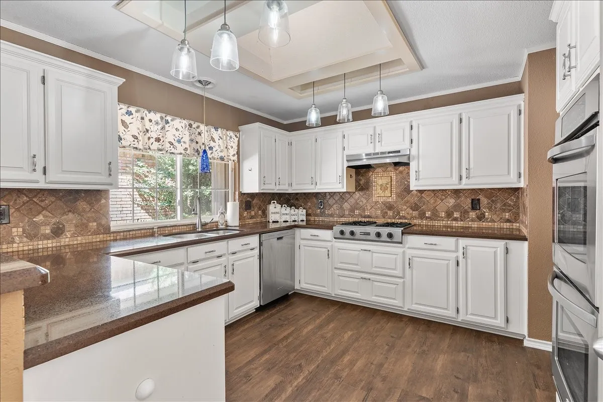 White cabinetry with stainless steel hardware, brown countertops, patterned tile backsplash, wood-finish flooring, and pendant lighting