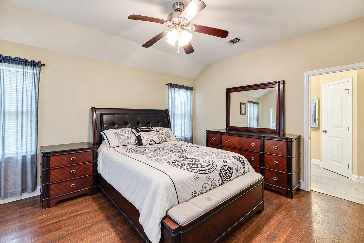 Vaulted ceiling featuring a ceiling fan with integrated lighting, wood-finish flooring, two windows with blinds, and an open doorway leading to another area with tile flooring
