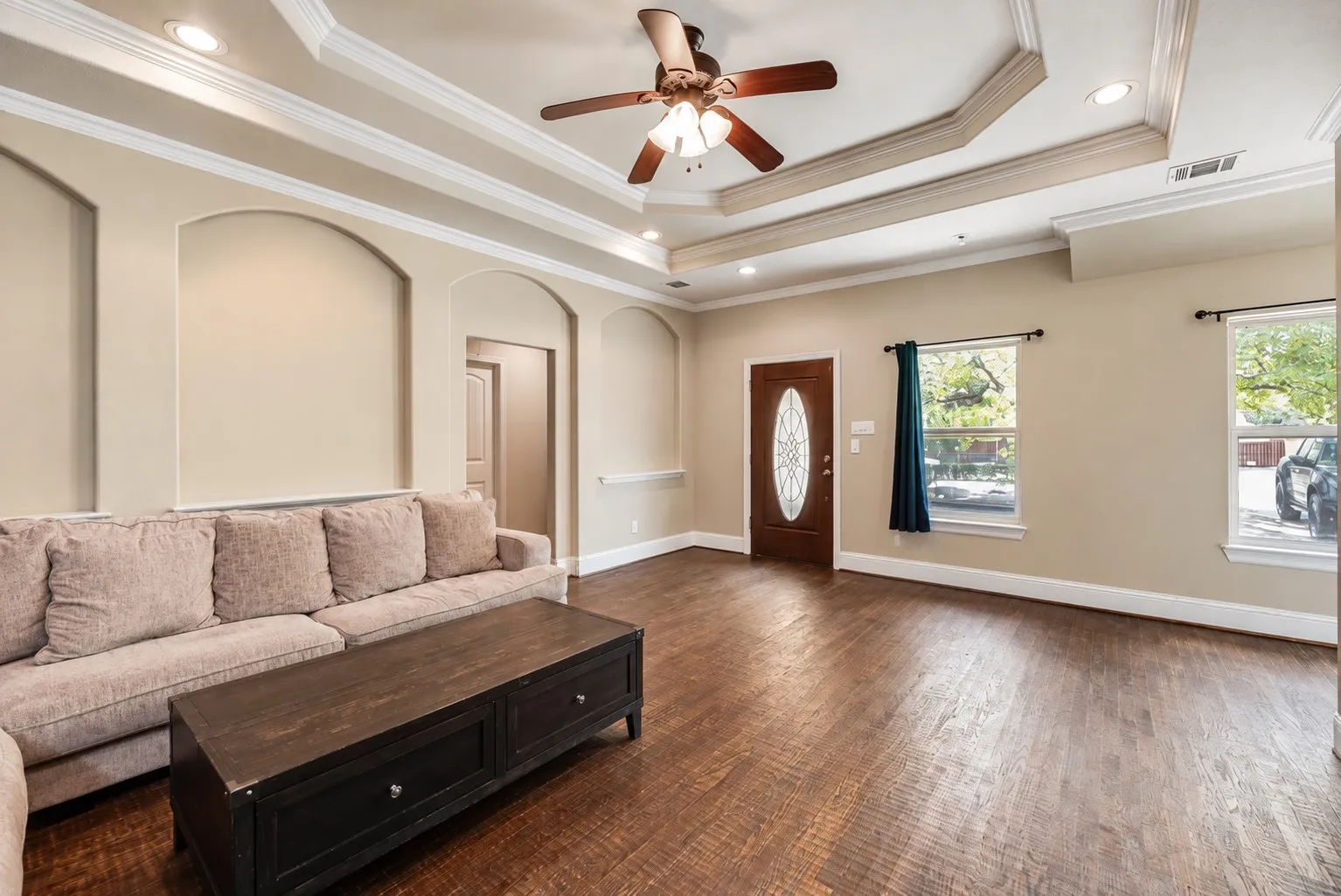 Wood-finish flooring, tray ceiling with recessed lighting, ceiling fan, arched wall cutouts, and a front entry door with an oval glass insert