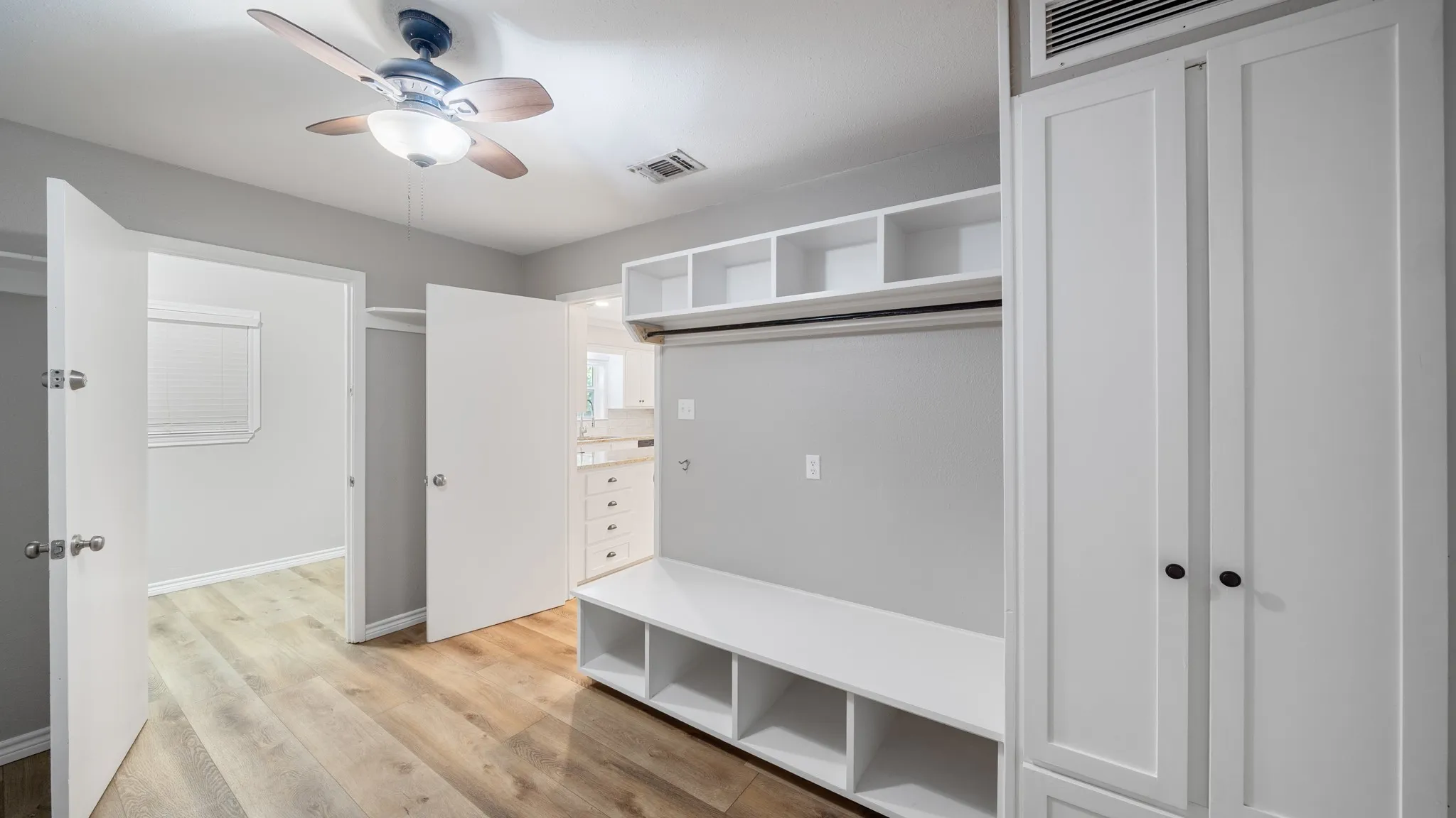 Wood-finish flooring, built-in storage bench, integrated shelving, white shaker-style cabinetry, and ceiling fan with integrated lighting