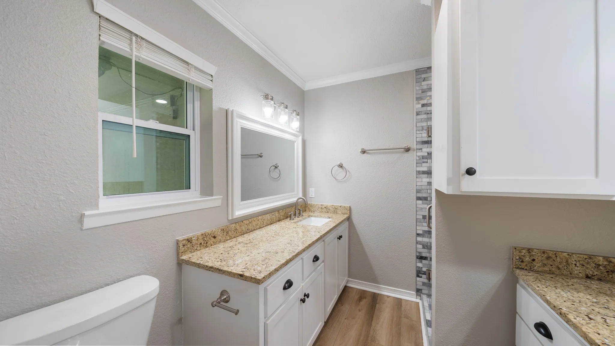 Double vanity with granite countertops, wood-finish flooring, white cabinetry, and a tiled shower surround