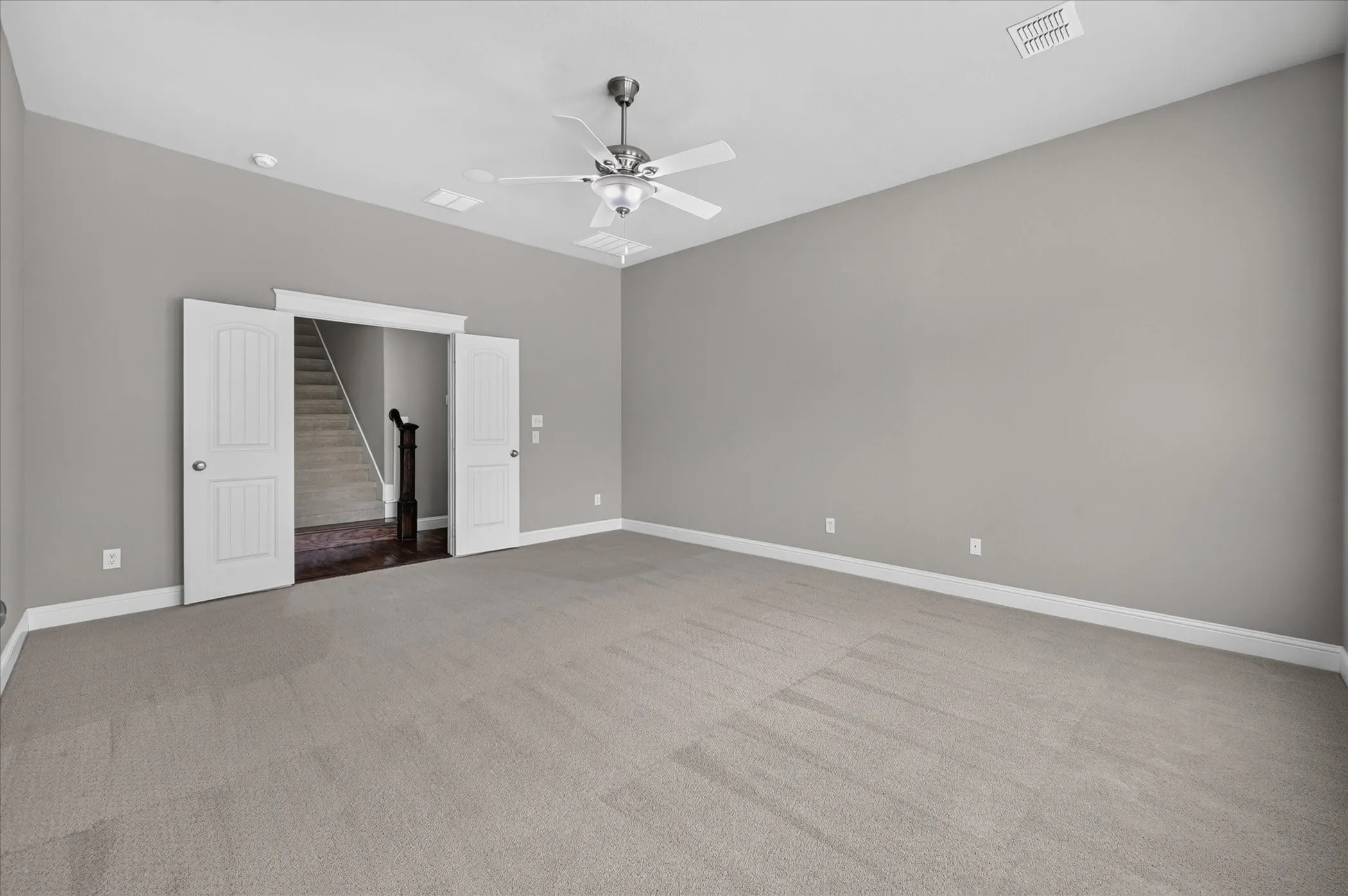 Expansive carpeted room featuring a ceiling fan with integrated lighting, a vaulted ceiling, and double doors opening to a staircase with a dark wood-finish newel post