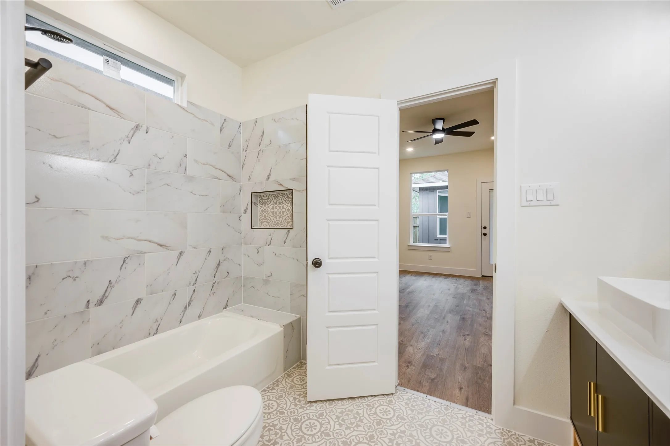 Tiled tub-shower combination with recessed niche, patterned floor tiles, modern vanity with brass hardware, and wood-finish flooring in the adjacent room