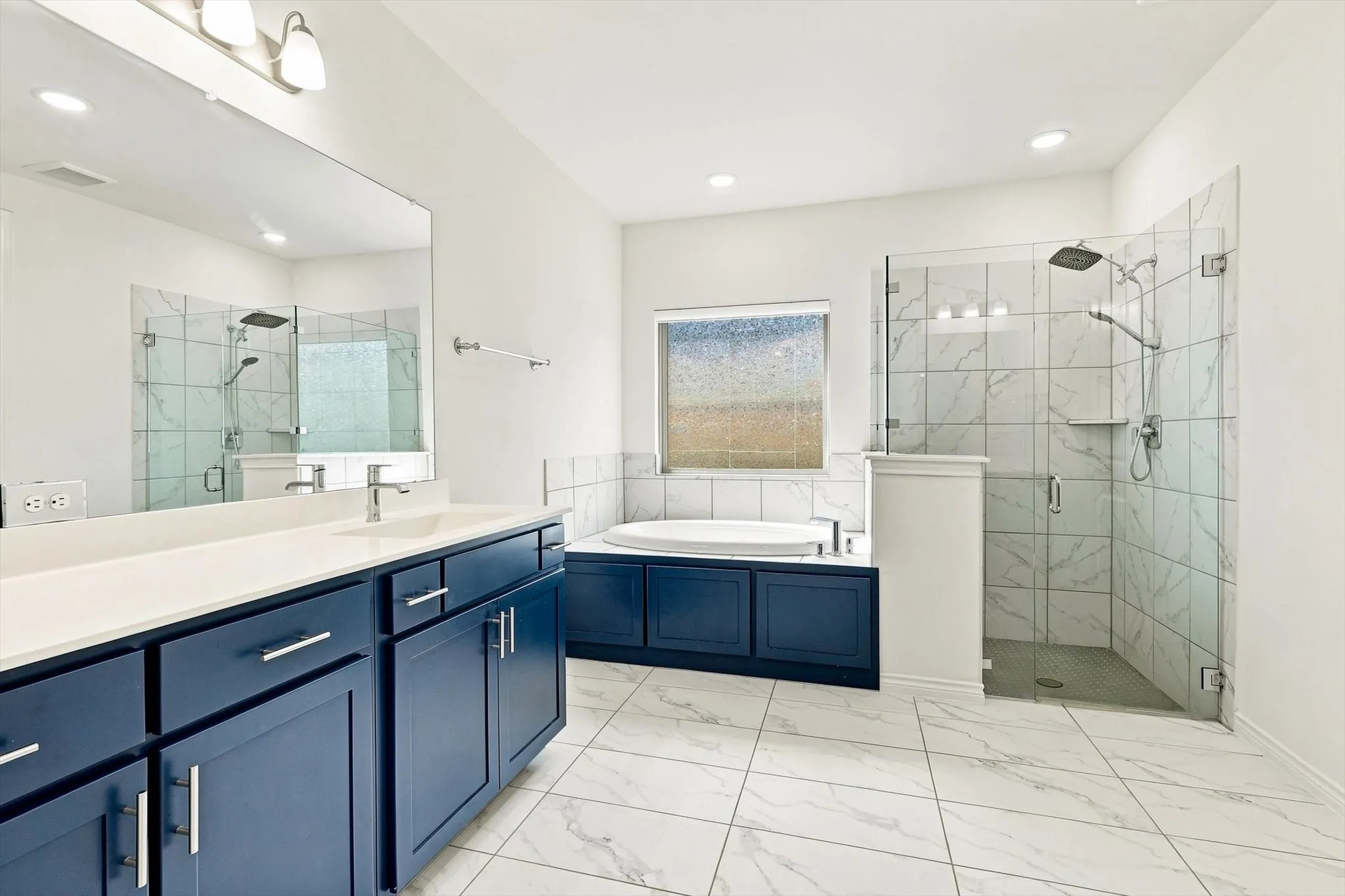 Dual vanity with blue cabinetry and white countertops, enclosed glass shower with tiled walls and rain showerhead, soaking tub with tiled surround, and white tile flooring