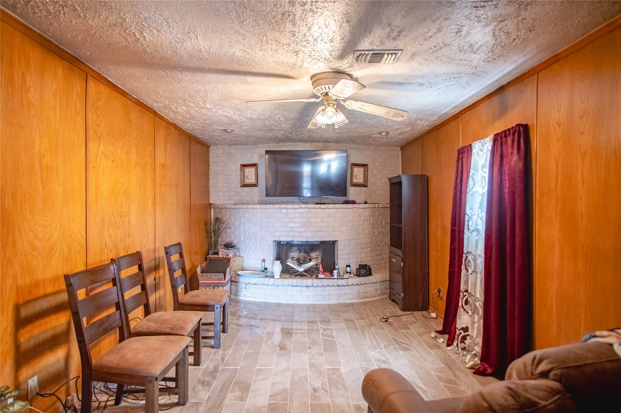 Wood-finish paneling, brick fireplace, textured ceiling, tiled flooring, ceiling fan with light fixture