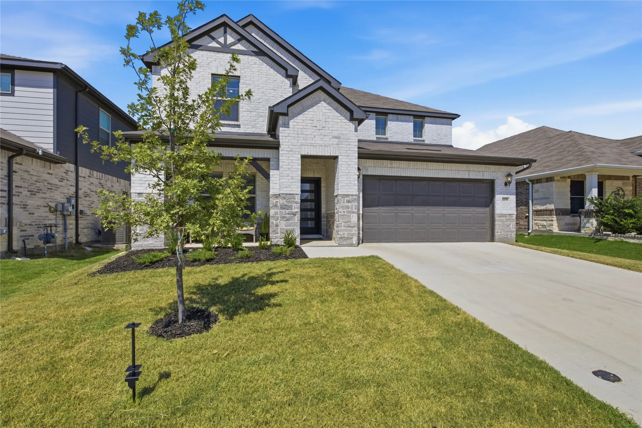 Contemporary brick and stone exterior with dark trim accents, concrete driveway, attached garage, and mature landscaping