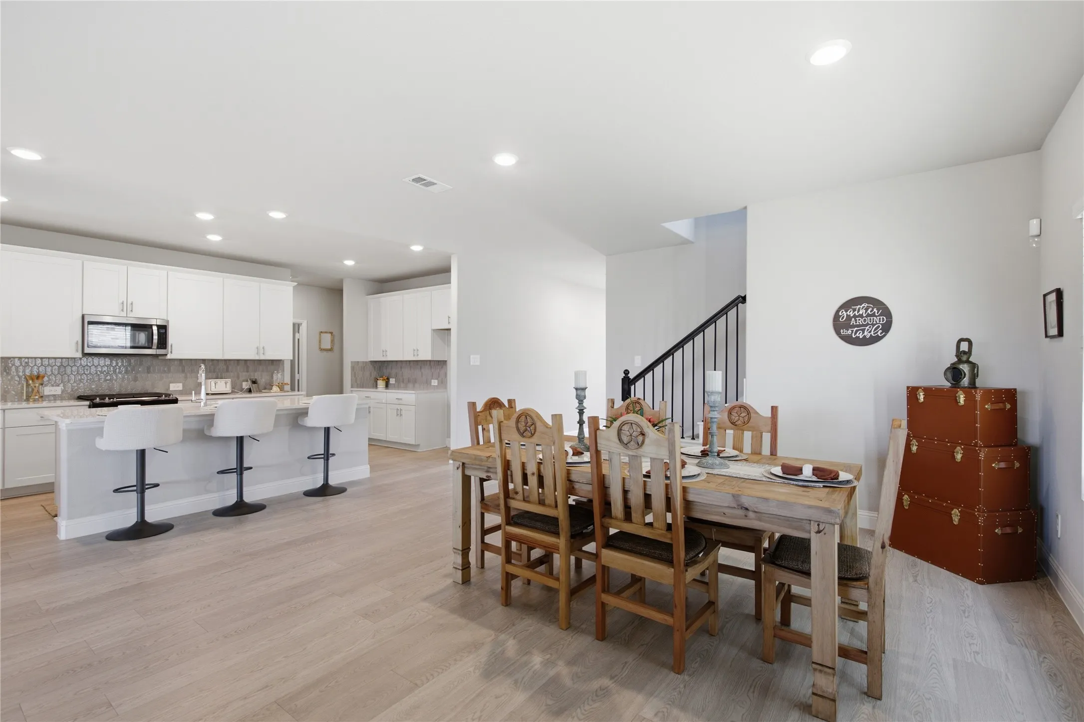 Open-concept living area featuring wood-finish flooring, a kitchen with white cabinetry and an island, a dining area, and a staircase with metal balusters