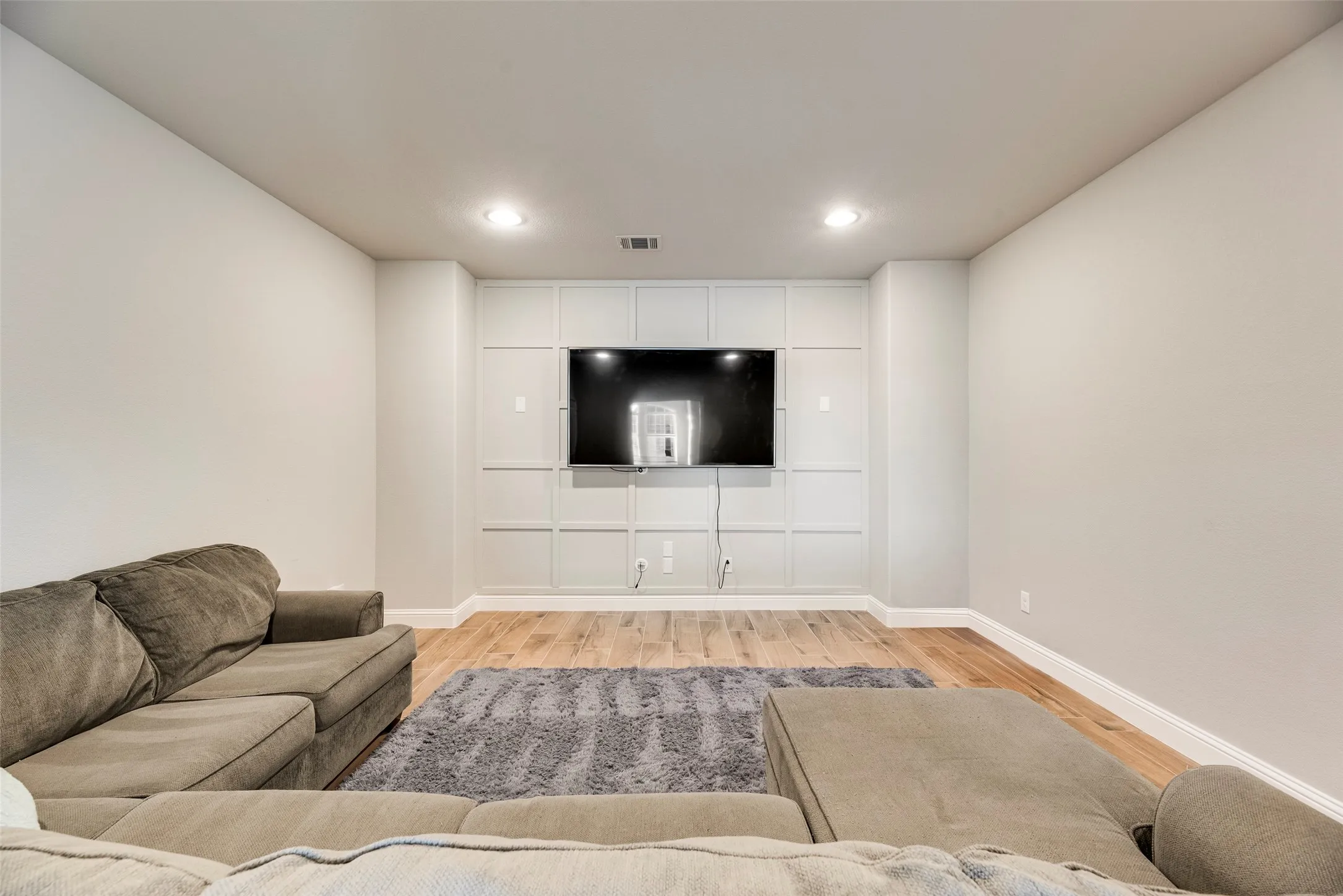 Recessed lighting illuminates the room, featuring wood-finish flooring and a built-in entertainment wall with panel detailing