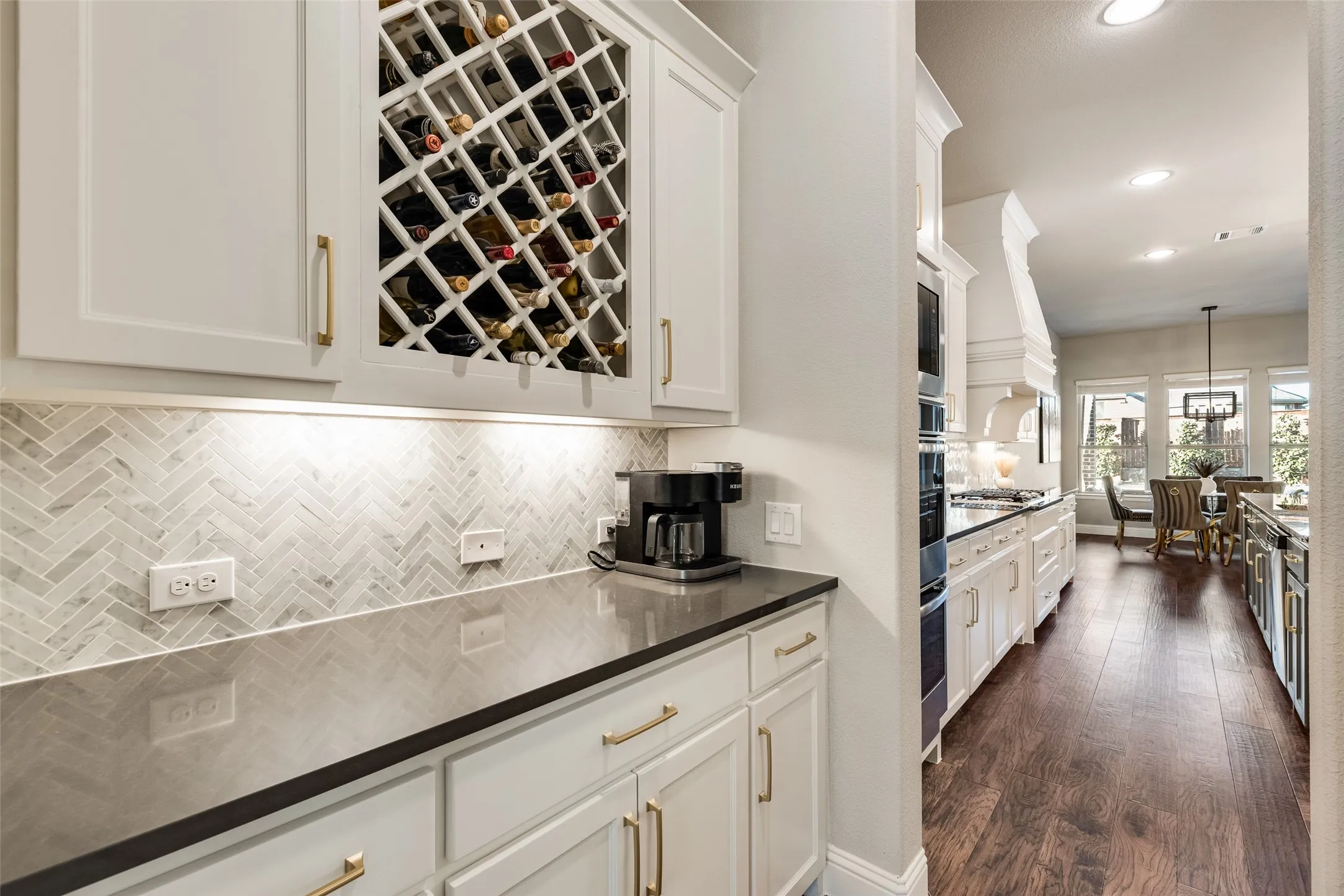 Built-in wine storage, white cabinetry with gold-tone hardware, dark countertops, herringbone tile backsplash, and recessed lighting