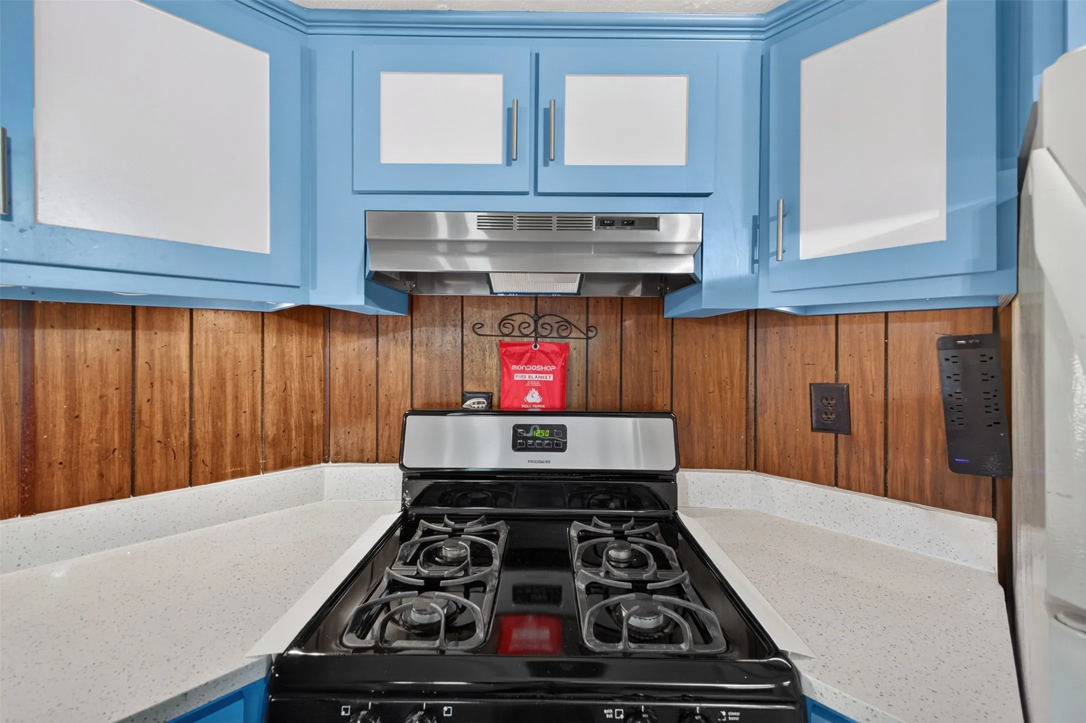 Kitchen featuring light blue upper cabinetry, wood panel backsplash, light-toned countertops, and a built-in range with an overhead stainless steel range hood