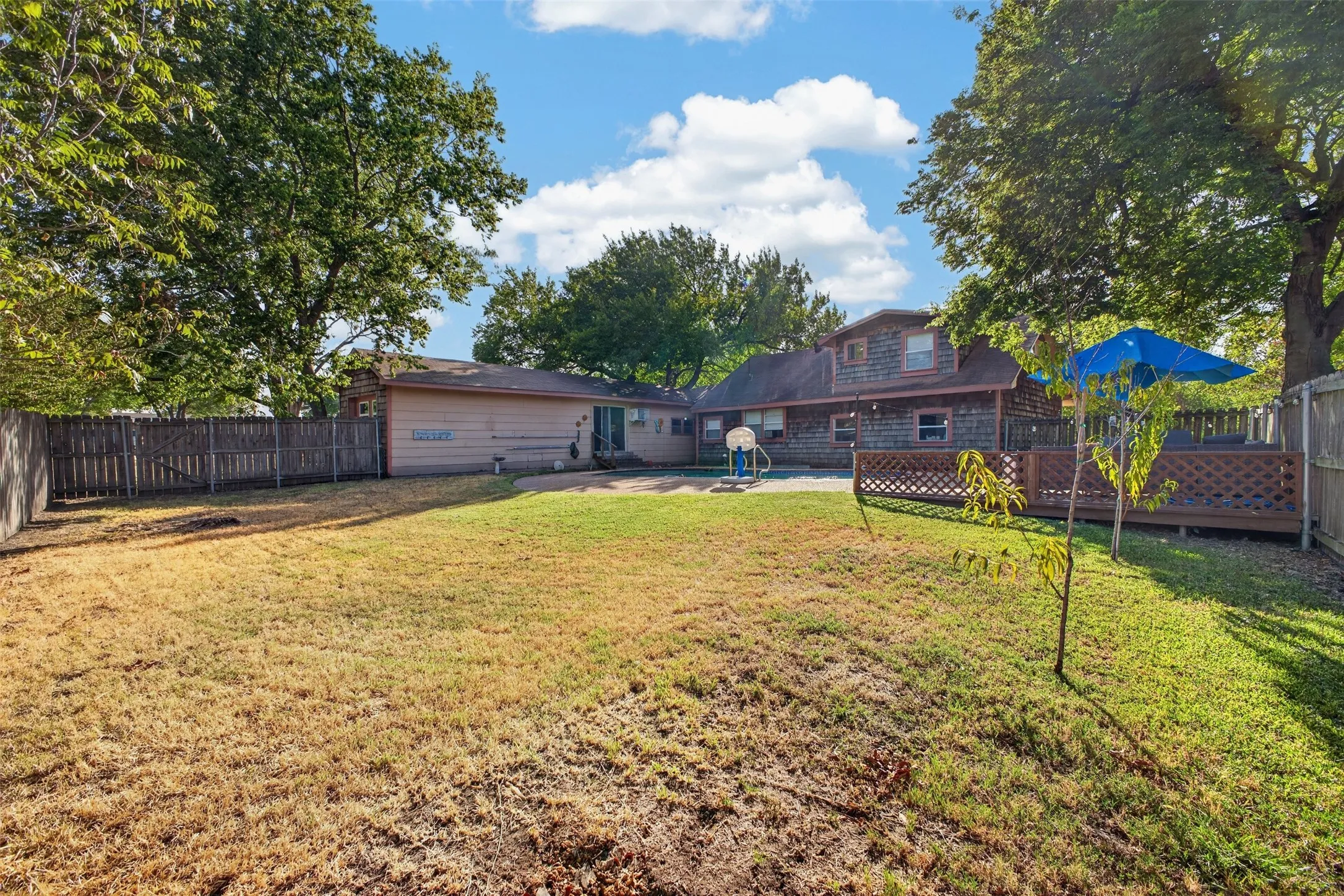 Expansive fenced backyard featuring a wooden deck with lattice railing, mature trees, and a paved patio area