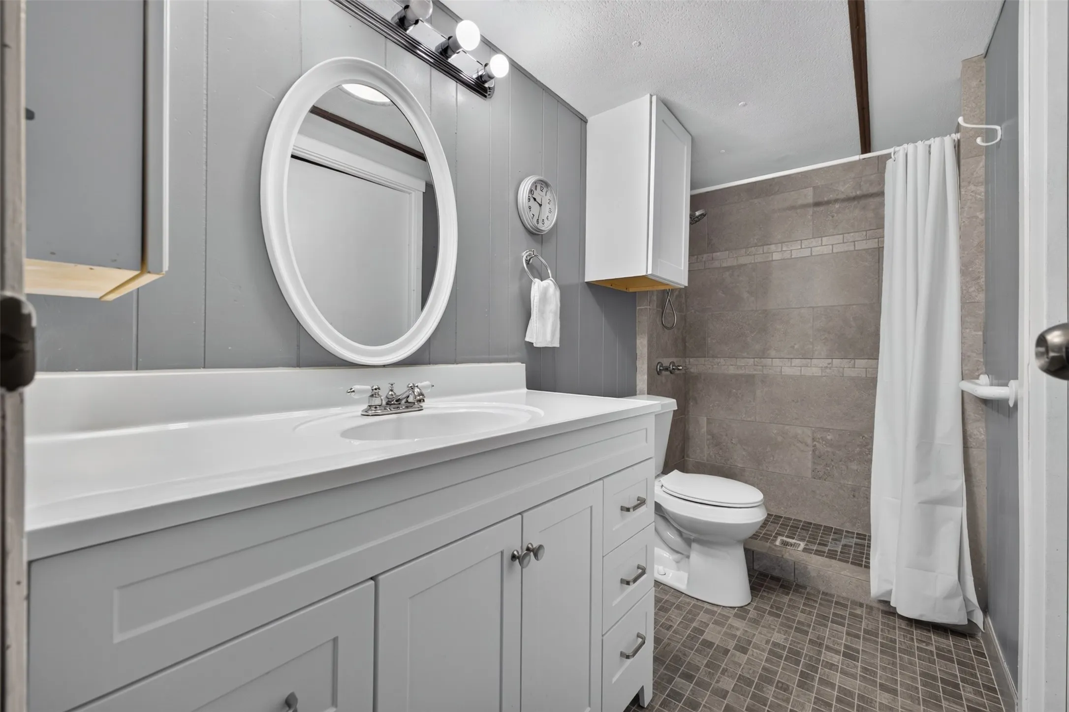 Vanity with a white countertop and integrated sink, gray wall paneling, oval mirror, gray tiled shower walls, and gray mosaic tile flooring