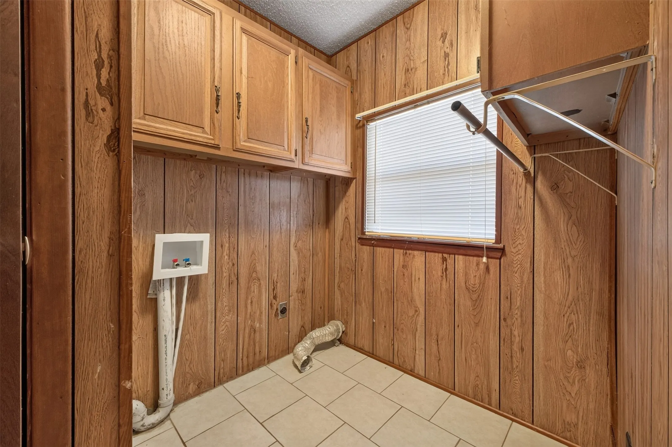 Integrated laundry area featuring wood-paneled walls, overhead cabinetry, a window with blinds, and tile flooring