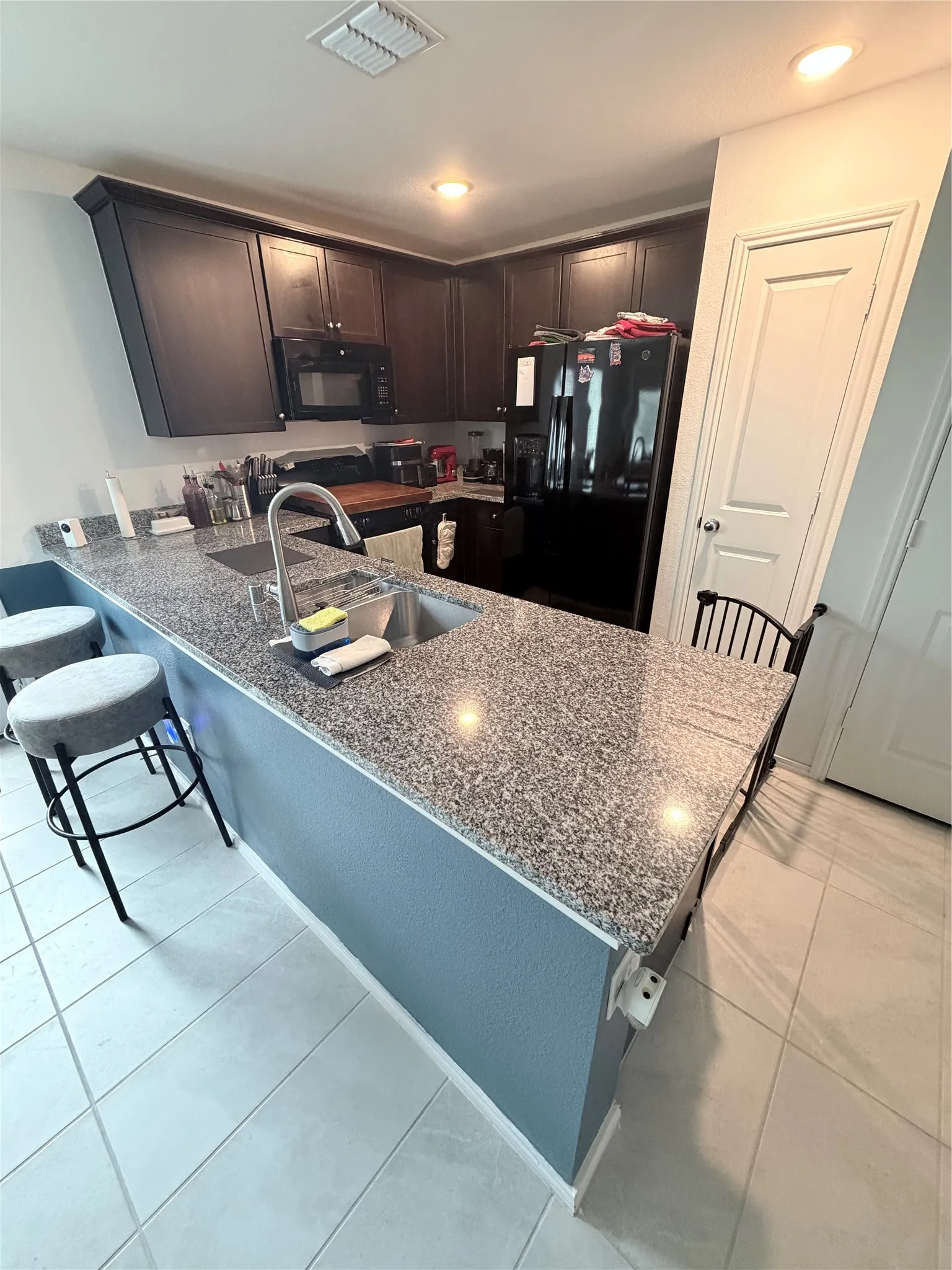 Kitchen featuring granite countertops, dark wood-finish cabinetry, a built-in microwave, a stainless steel sink with a high-arc faucet, and tiled flooring