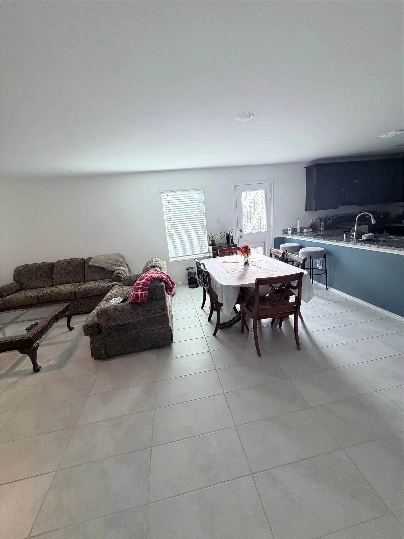 Open-concept living space featuring light-colored tile flooring, recessed lighting, a window with blinds, a modern kitchen island with seating, and dark cabinetry
