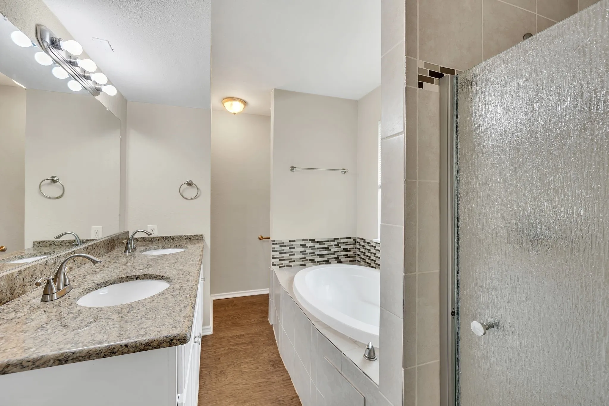 Unit C- Dual vanity with oval undermount sinks and brushed nickel faucets, granite-style countertop, large mirror, drop-in soaking tub with tile surround, and an enclosed shower with textured glass