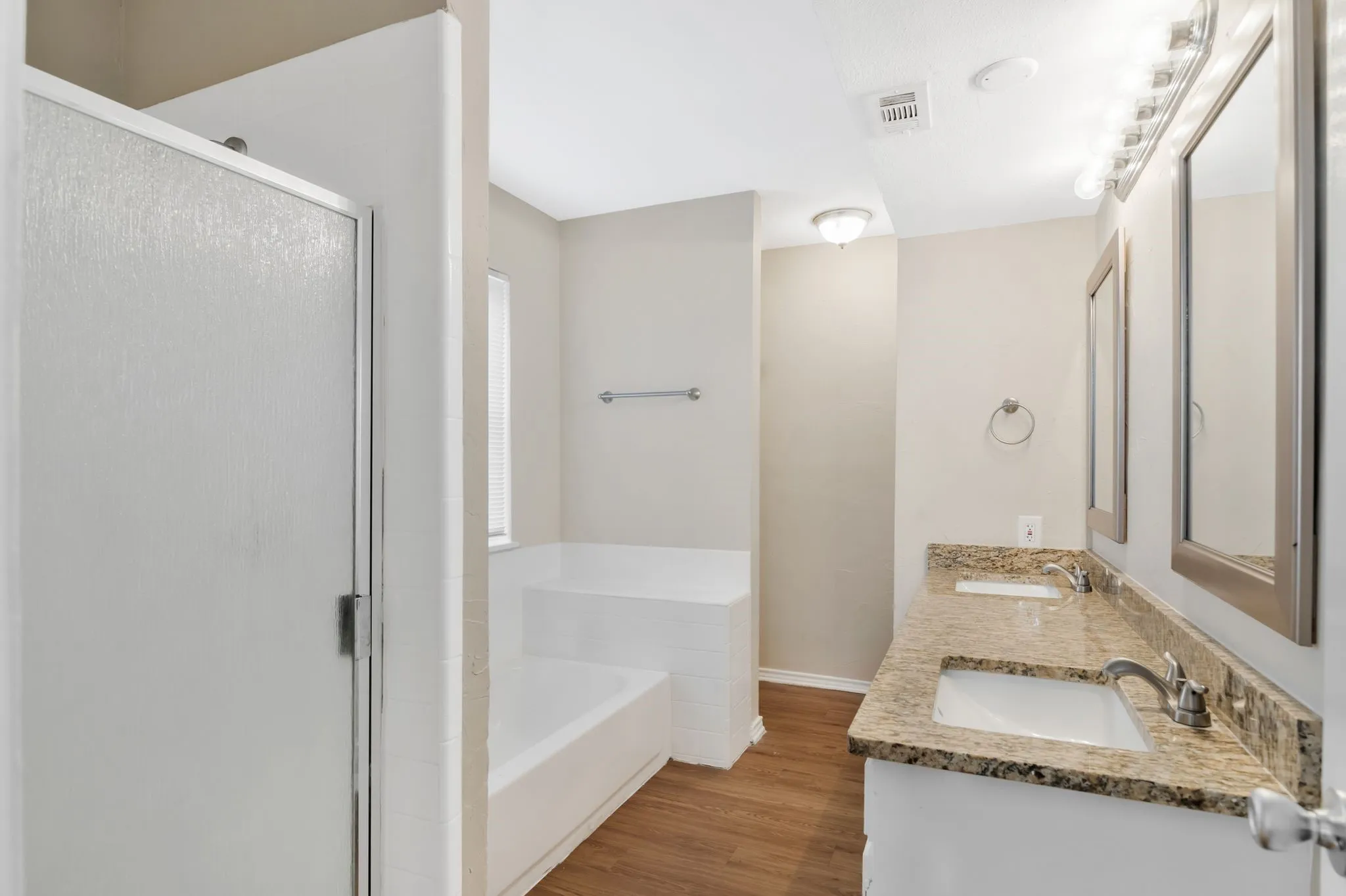Unit B -Dual vanity with granite countertops, integrated sinks, brushed nickel fixtures, and individual framed mirrors, complemented by a separate shower with a frosted glass door and a built-in tub