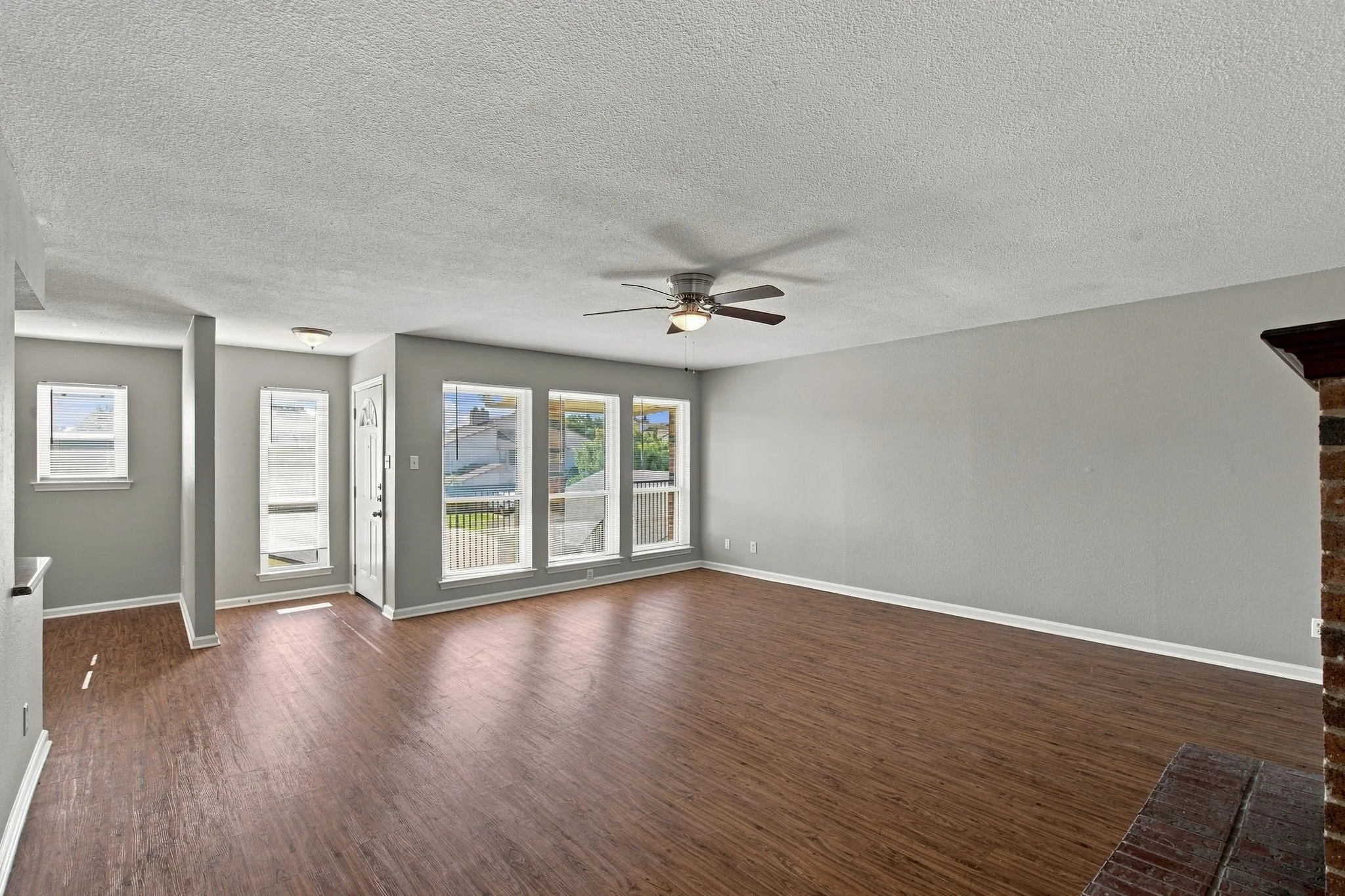 Unit D -Spacious living area featuring wood-finish flooring, light gray wall paint, multiple large windows, and a ceiling fan